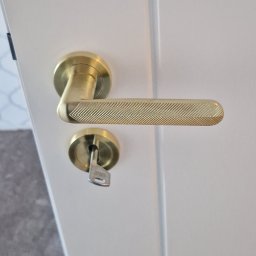 Grzegorz Sadło - Białe drzwi z złotą klamką o wzorze jodełki i kluczem w zamku, widok z bliska. Detal wykończenia wnętrza.