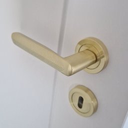 Grzegorz Sadło - Białe drzwi z zamontowaną złotą klamką o geometrycznym wzorze, widoczne z bliska, z delikatnym oświetleniem podkreślającym detale wykończenia.