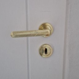 Grzegorz Sadło - Białe drzwi z zamontowaną, złotą klamką o prążkowanej fakturze i szyldem zamka. Minimalistyczny design, subtelne oświetlenie.