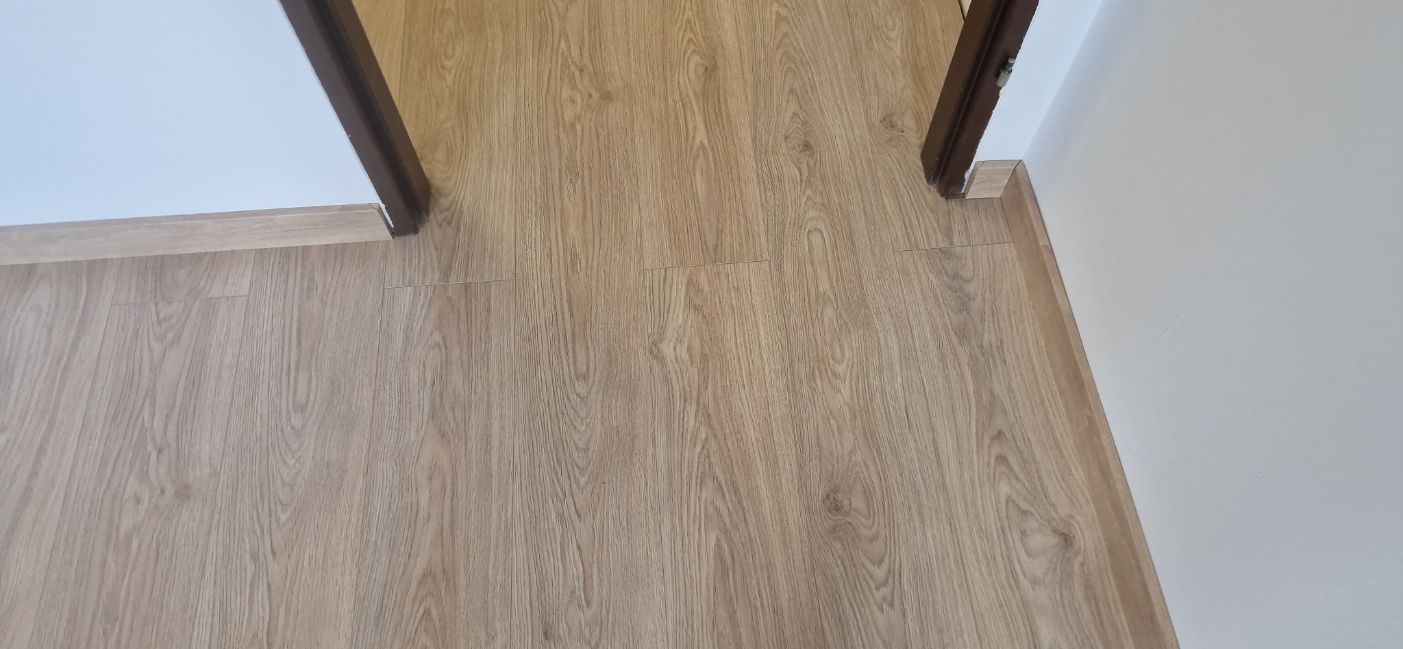 Jasne panele podłogowe ułożone wzdłuż ściany i progu drzwiowego. Widoczne listwy przypodłogowe w kolorze paneli. Ujęcie z góry, naturalne światło.