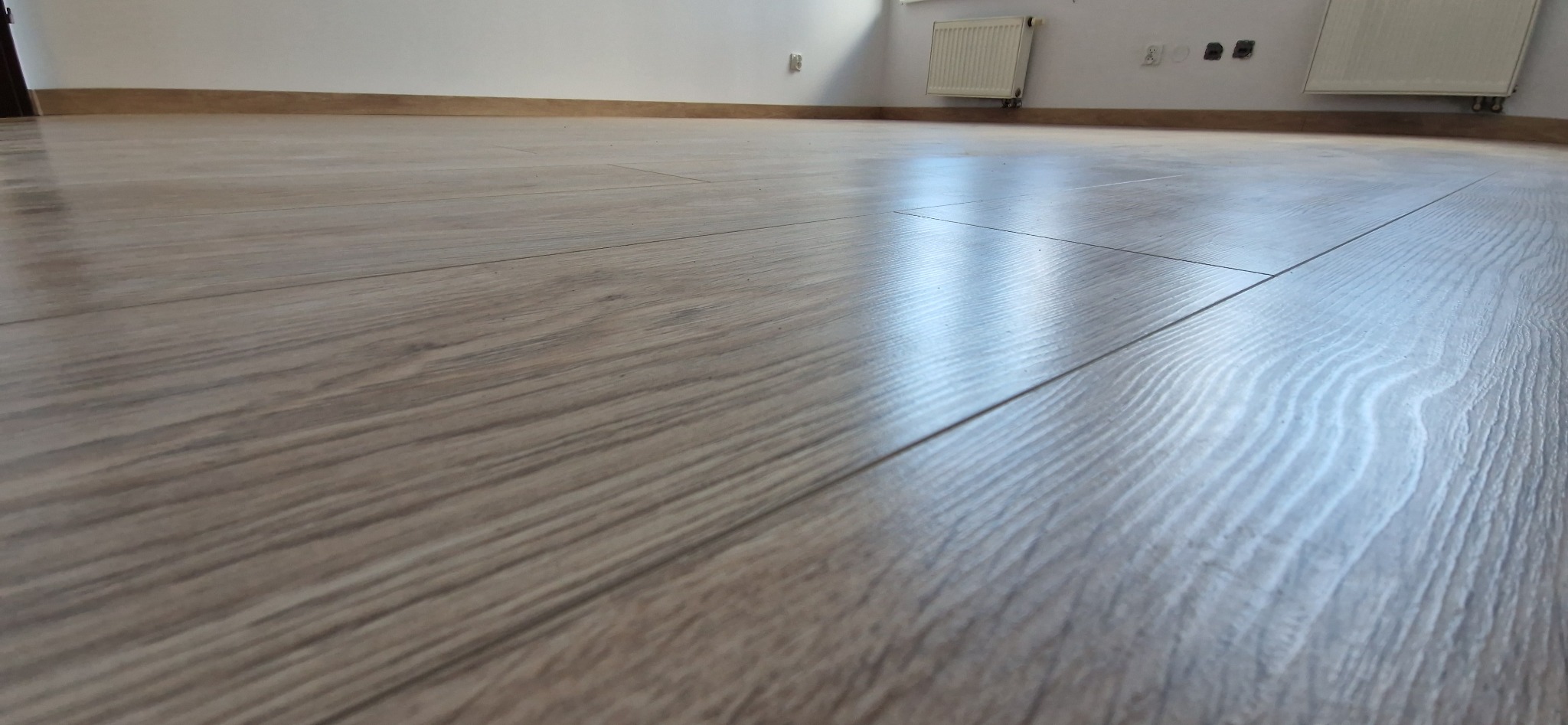 Jasne panele podłogowe z widoczną strukturą drewna, ułożone w nowym wnętrzu. W tle biała ściana z grzejnikiem i listwą przypodłogową.