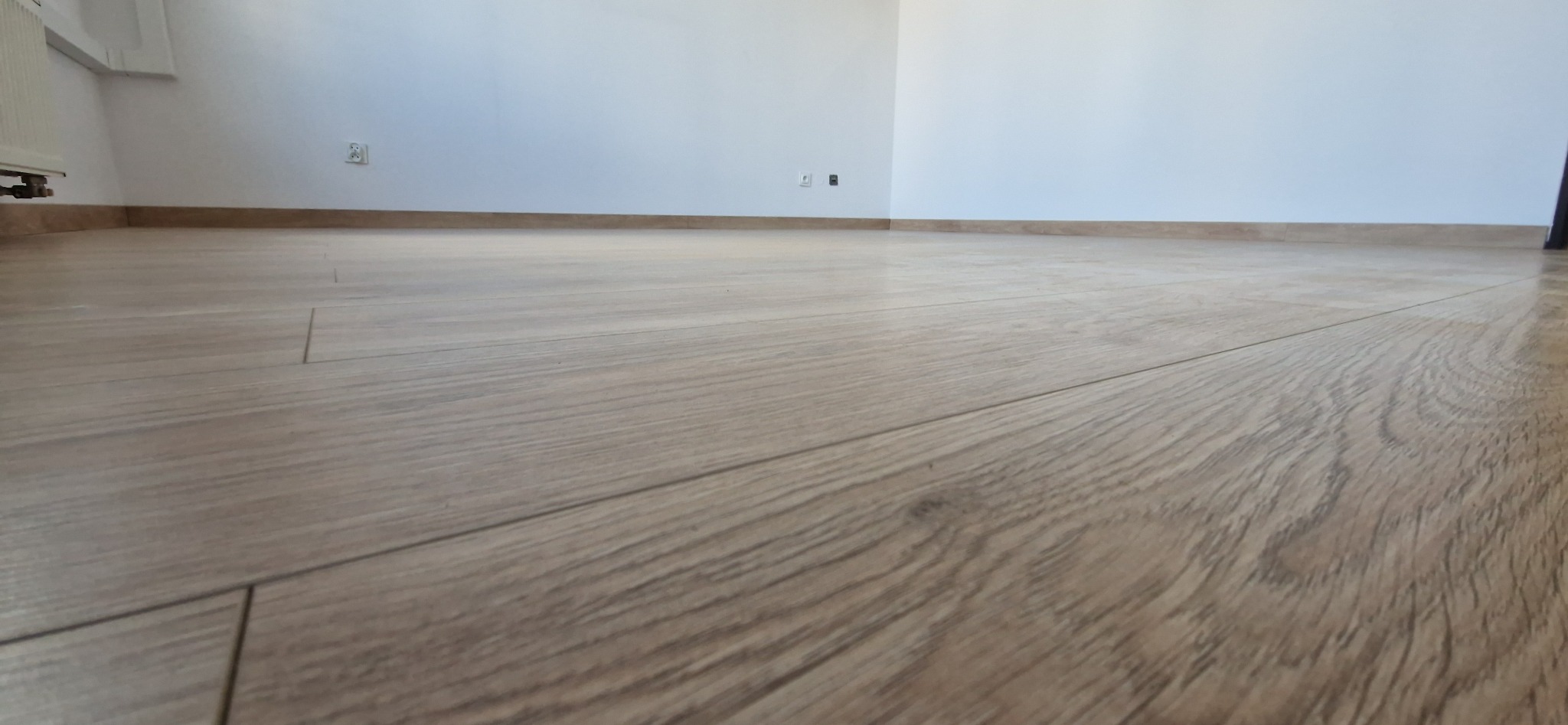 Jasne panele podłogowe ułożone w pokoju z białymi ścianami i brązowymi listwami przypodłogowymi. Widoczne gniazdka elektryczne. Ujęcie z niskiej perspektywy.