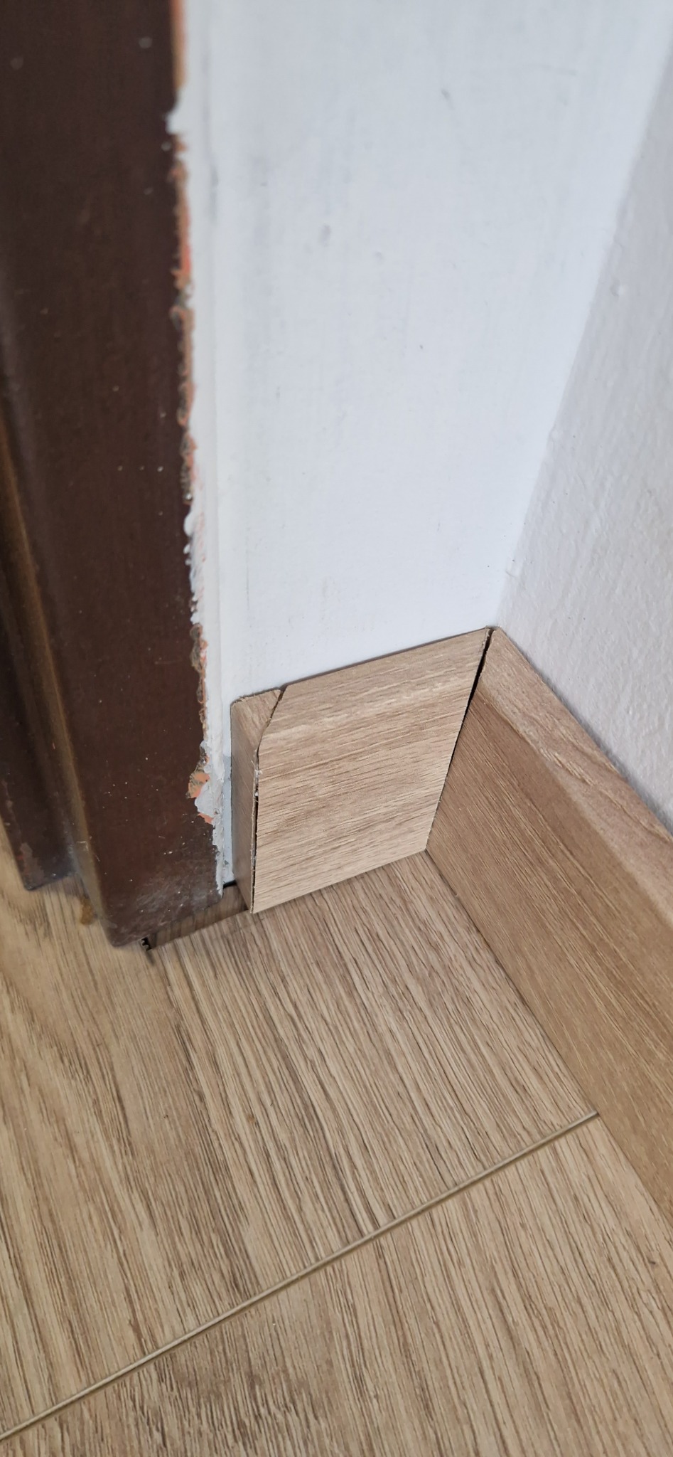 Detal wykończenia podłogi: brązowe panele podłogowe i listwa przypodłogowa z imitacją drewna, przy brązowej futrynie drzwi i białej ścianie. Widoczne niedoskonałości.