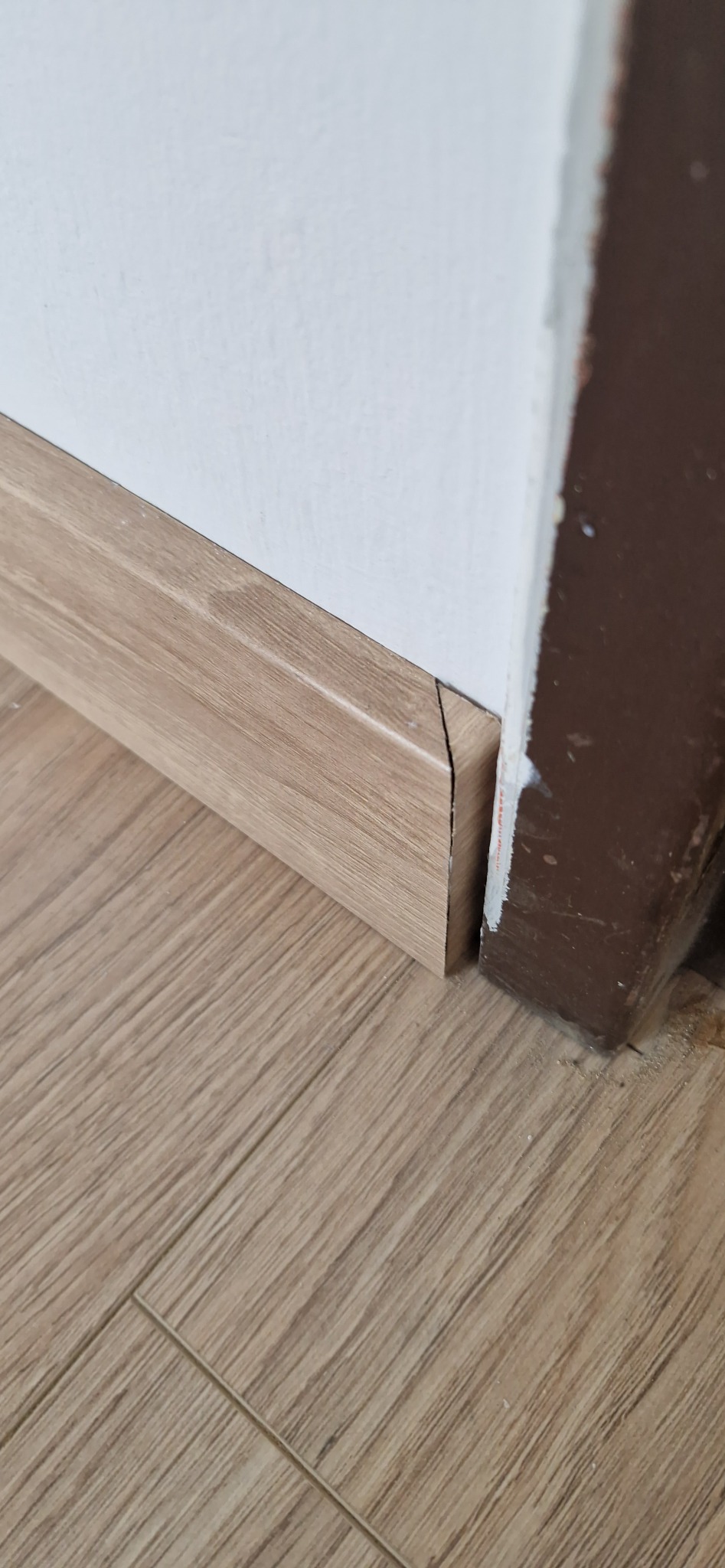 Fragment podłogi z paneli w odcieniach beżu, listwa przypodłogowa w kolorze drewna, stykająca się z brązową futryną drzwi. Widoczne niedoskonałości w wykończeniu.