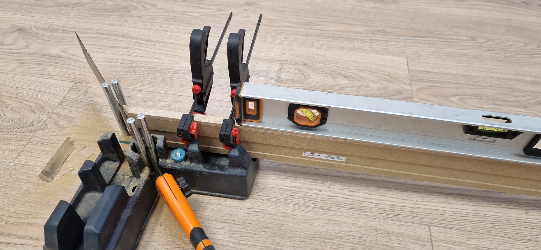 Przygotowanie do cięcia listwy przypodłogowej w skrzynce uciosowej, poziomica i ściski stolarskie na podłodze imitującej drewno. Narzędzia pracy.