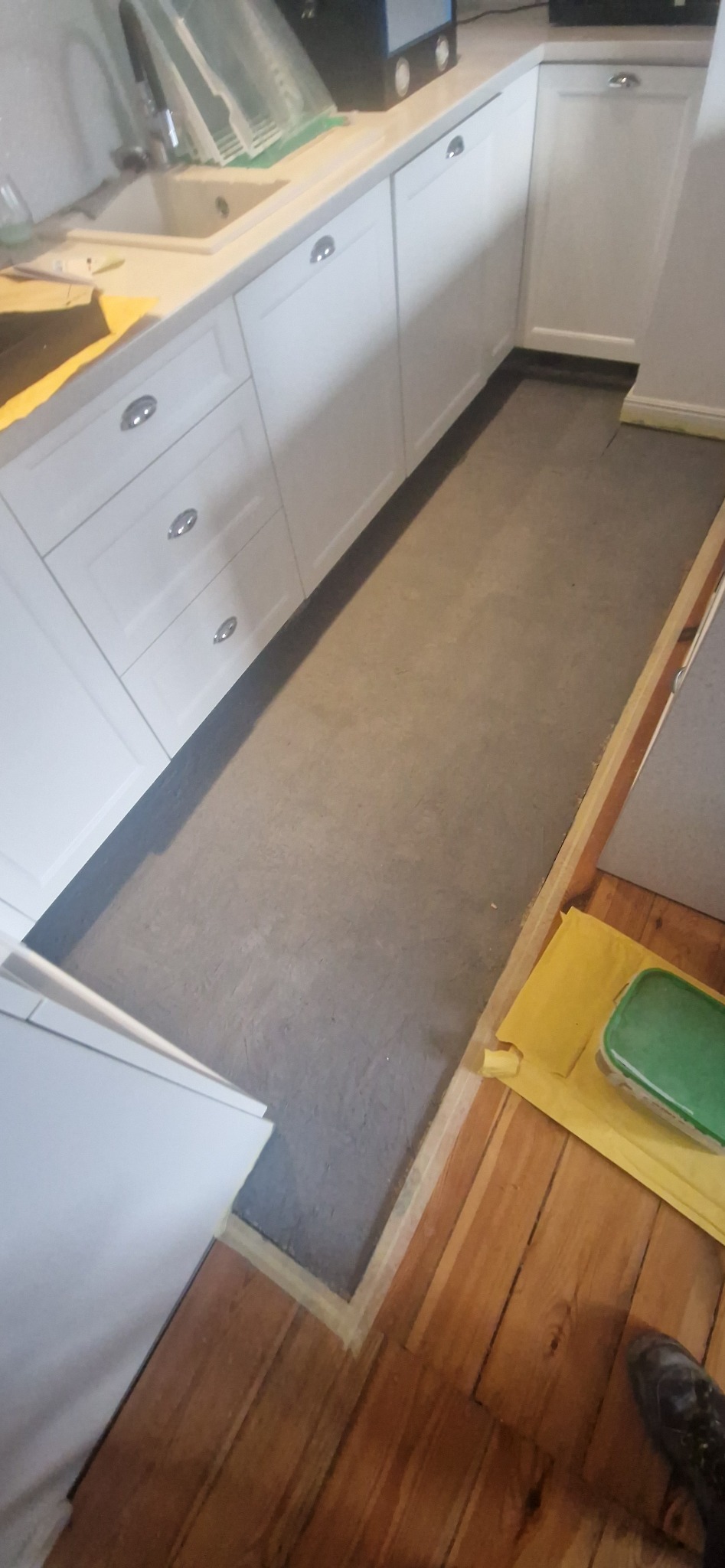 Przygotowanie do układania paneli w kuchni: widoczna wylewka podłogowa, białe szafki kuchenne i drewniana podłoga zabezpieczona folią. Etap prac wykończeniowych.