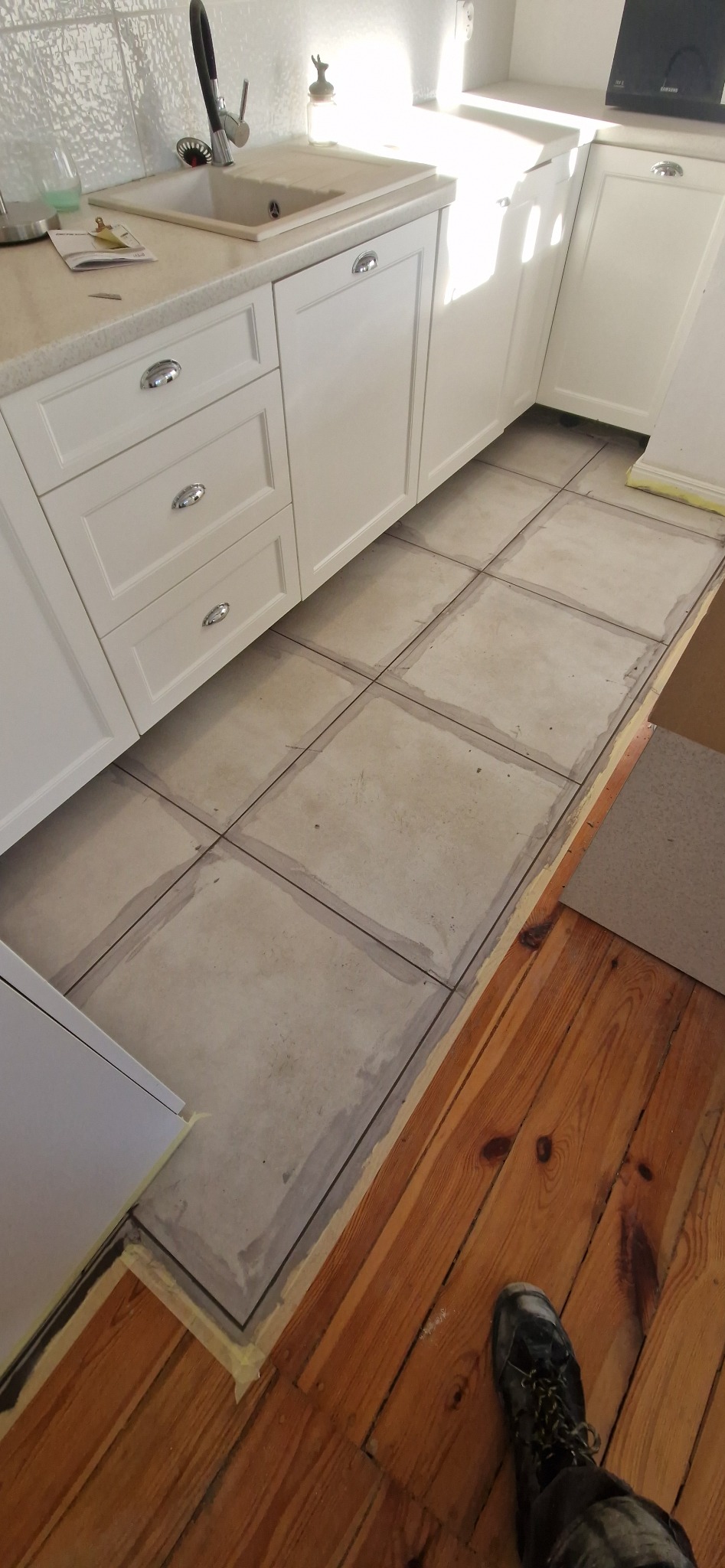 Kuchnia w trakcie remontu: białe szafki kuchenne, szare płytki podłogowe z widocznymi fugami, fragment drewnianej podłogi i but roboczy w rogu kadru.