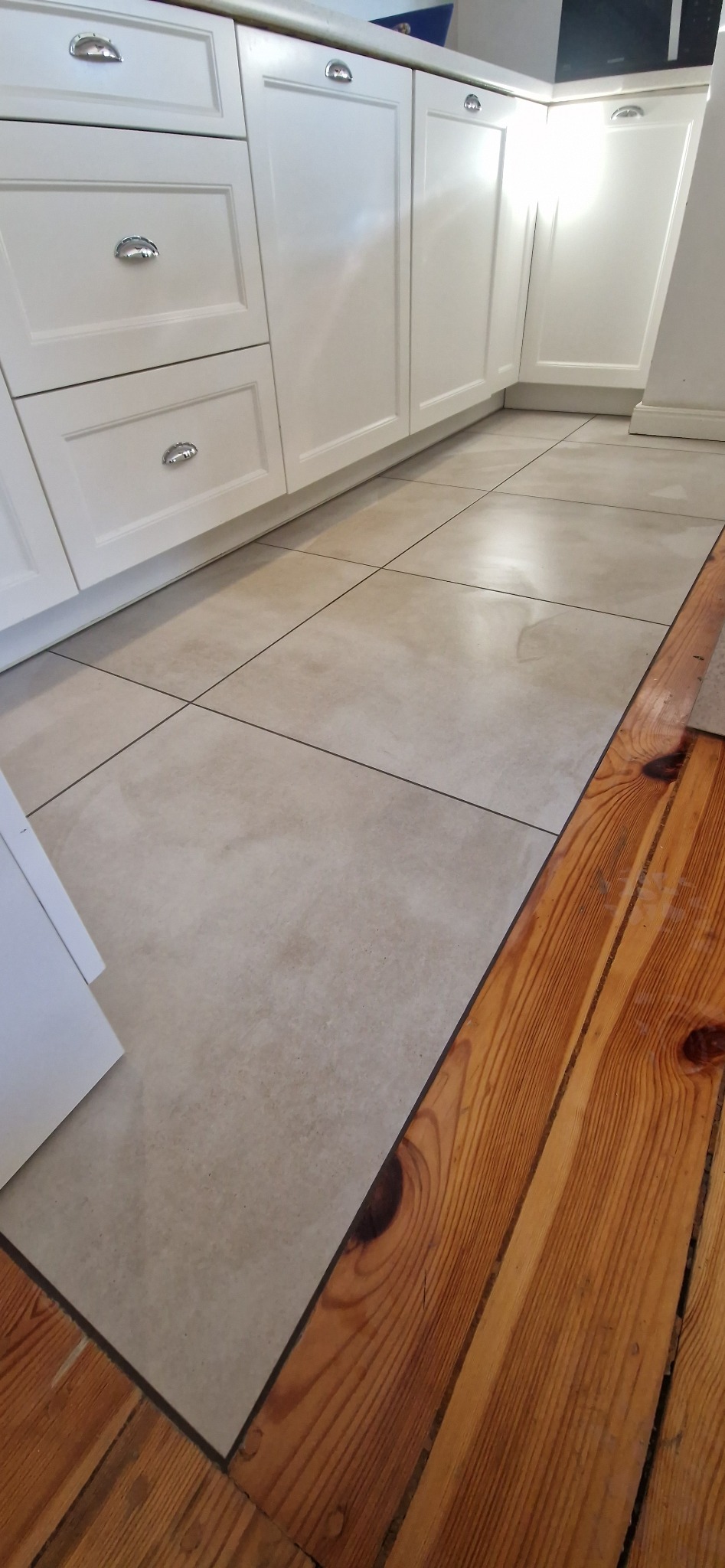 Beżowe płytki podłogowe w kuchni z białymi szafkami z metalowymi uchwytami, kontrastujące z fragmentem drewnianej podłogi. Widok z niskiej perspektywy.