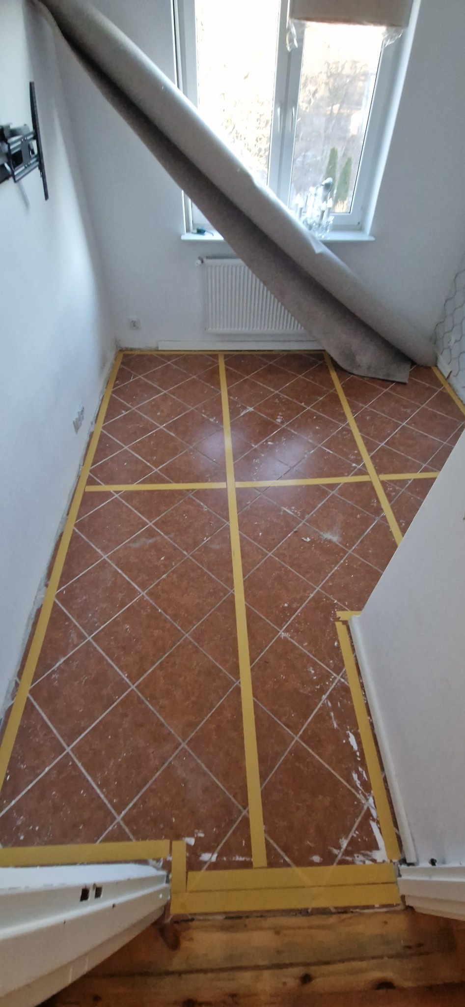 Przygotowanie do ułożenia paneli we Wrocławiu. Brązowe płytki podłogowe z wyznaczonym obszarem taśmą malarską. Częściowo zwinięty dywan przy oknie.