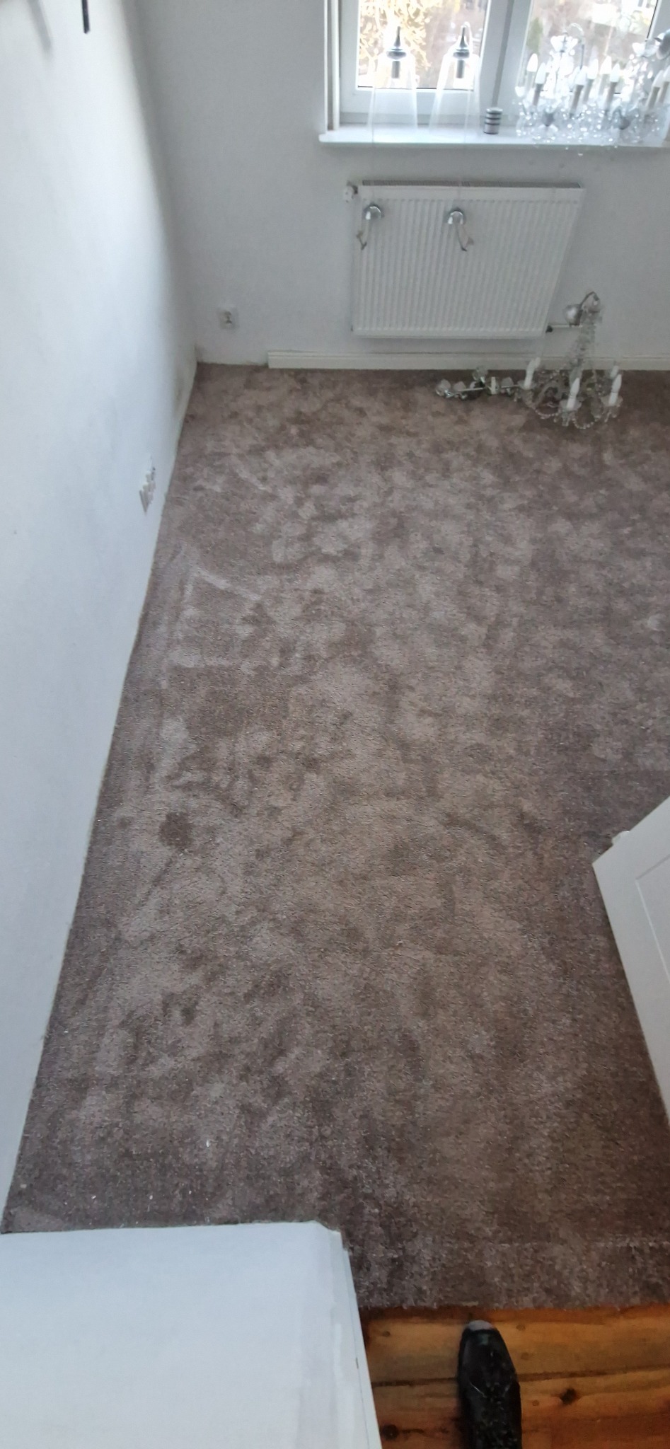 Pokój po położeniu brązowej wykładziny dywanowej. Widoczny fragment okna z dekoracjami, grzejnik i fragment żyrandola. Wykładzina o jednolitej strukturze i kolorze.