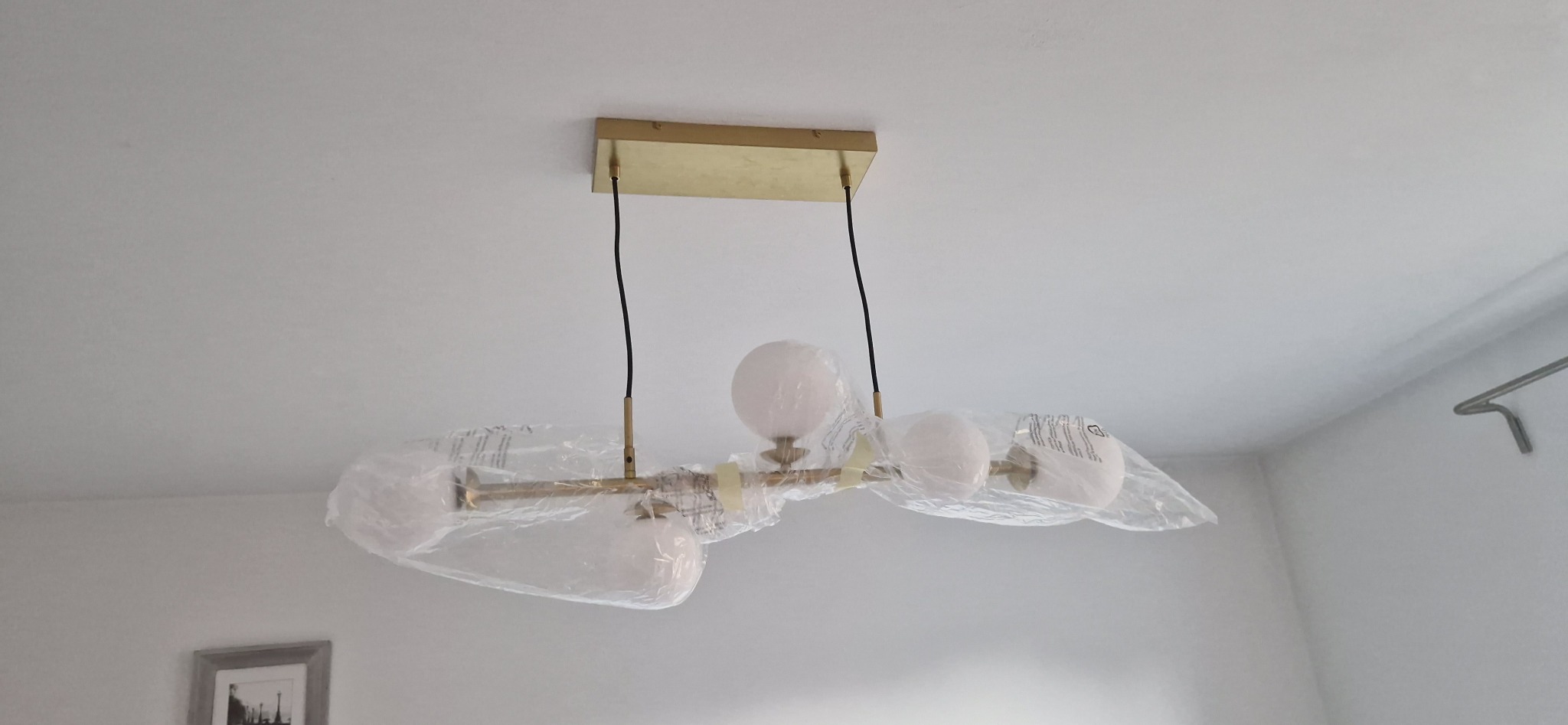 Nowoczesna, złota lampa sufitowa z trzema białymi kloszami w folii ochronnej, zamontowana na białym suficie. Widoczny fragment ściany i obrazek w ramce.