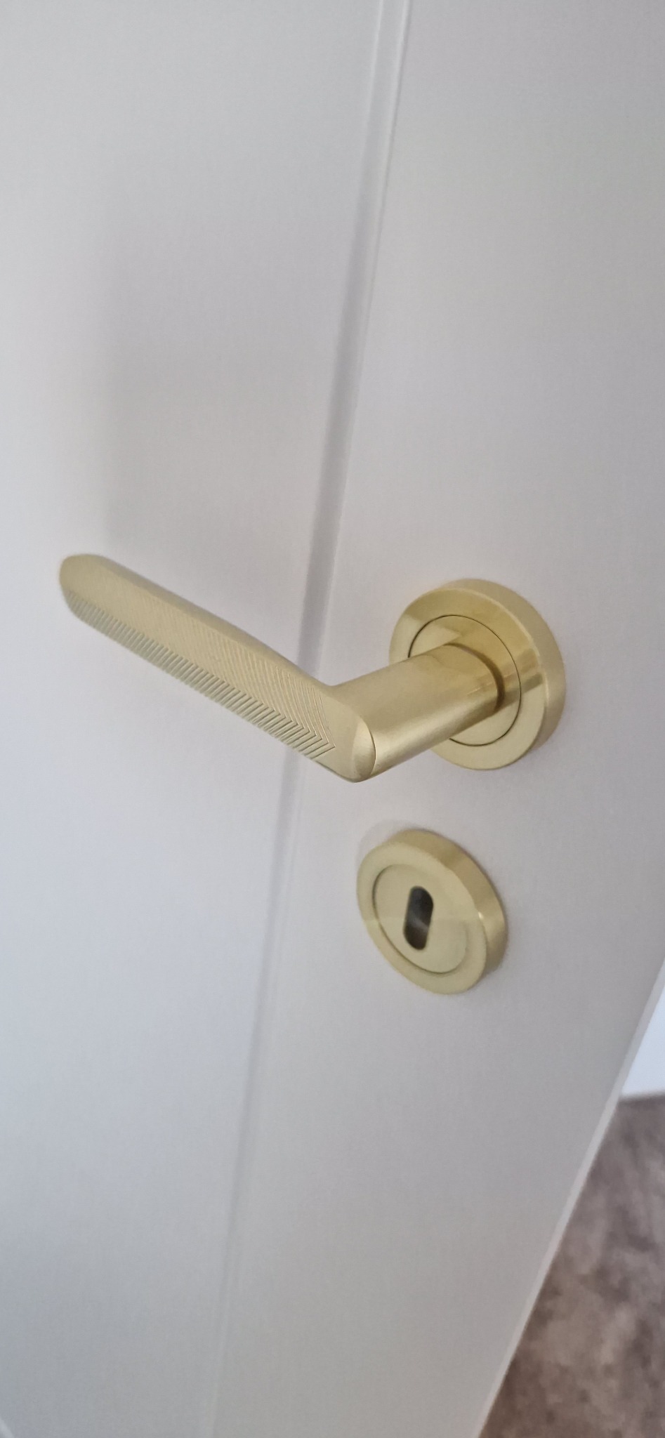 Białe drzwi z zamontowaną złotą klamką o geometrycznym wzorze, widoczne z bliska, z delikatnym oświetleniem podkreślającym detale wykończenia.