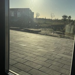 Michał - Ułożona, szara kostka brukowa widziana przez okno. W tle budynek w budowie i drzewa. Słoneczny dzień.