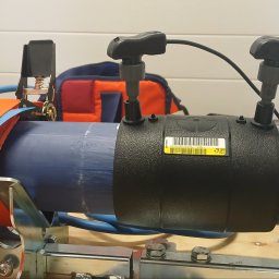 Michał - Zgrzewanie elektrooporowe rury PE z użyciem specjalistycznej zgrzewarki. Widoczne złącze, rura, pasy mocujące i narzędzia. Detal z bliska, oświetlenie warsztatowe.