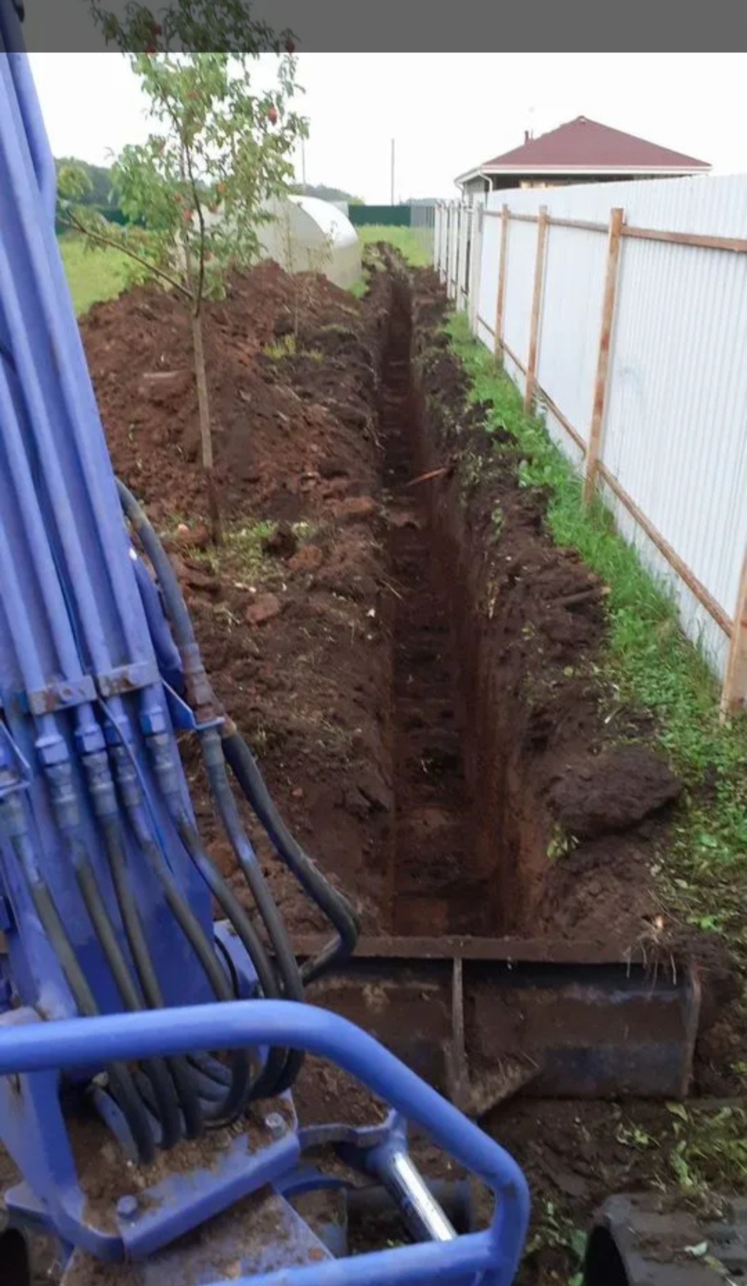 Wąski wykop w ziemi, wykonany prawdopodobnie pod instalację, widoczny z perspektywy operatora minikoparki, z fragmentem maszyny w kadrze i ogrodzeniem w tle.