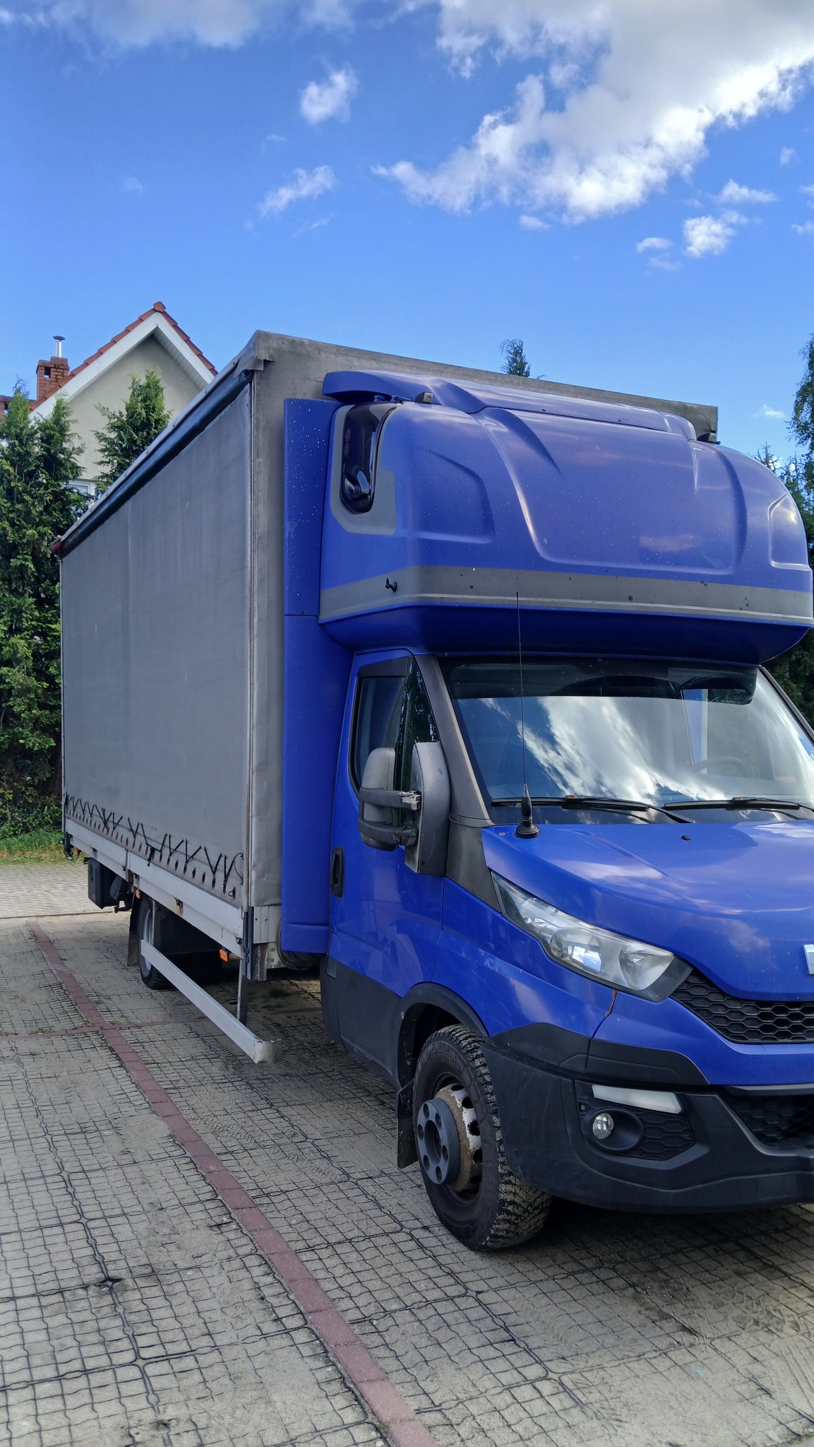 Niebieski samochód dostawczy Iveco z plandeką na parkingu w Rumi, z widocznymi detalami karoserii i opon. W tle budynek i drzewa.