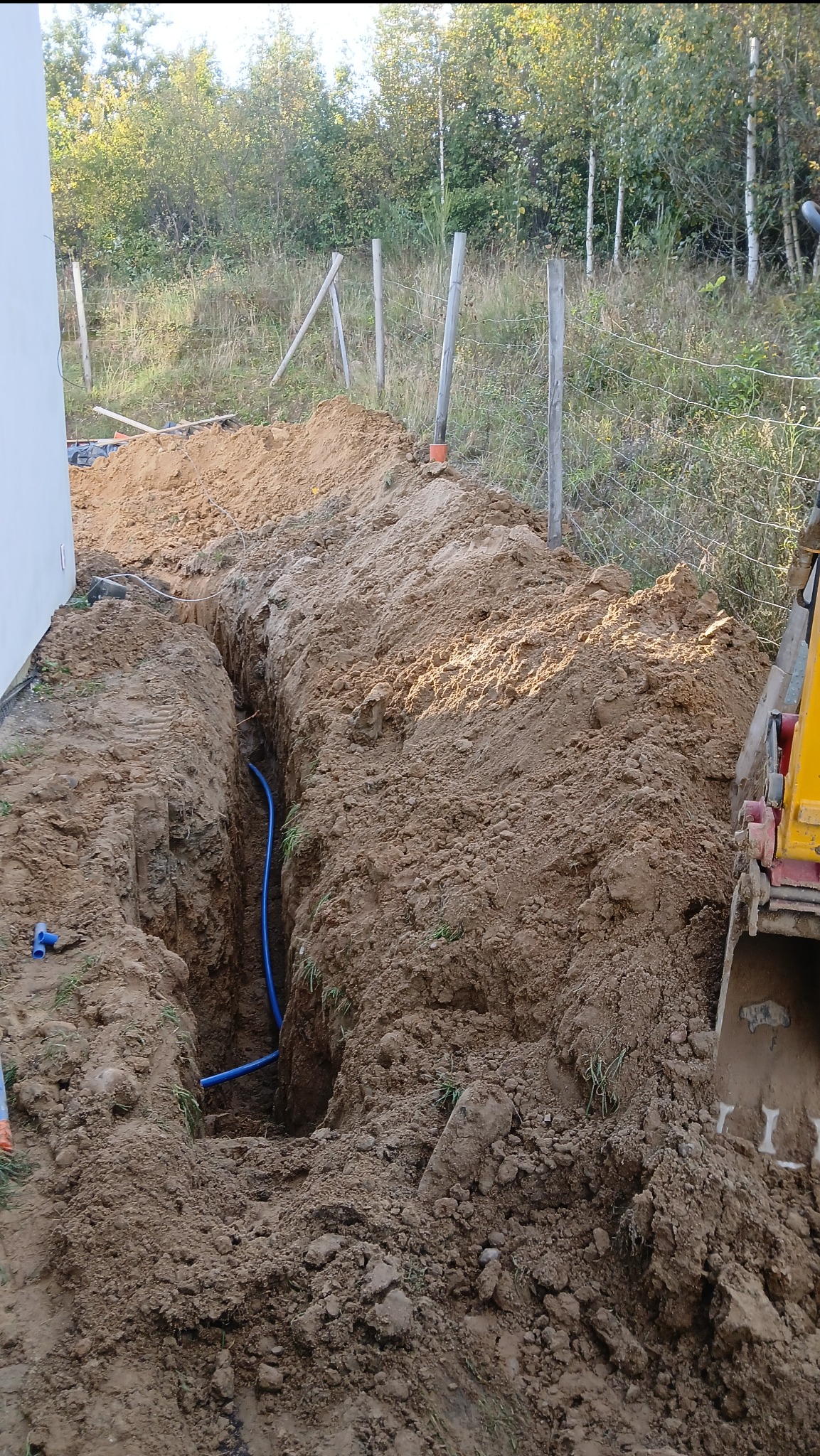 Wykopany rów z niebieskimi rurami w ziemi, widoczny fragment łyżki koparki. Tło: trawa, drzewa i drewniane ogrodzenie. Dzień, naturalne światło.