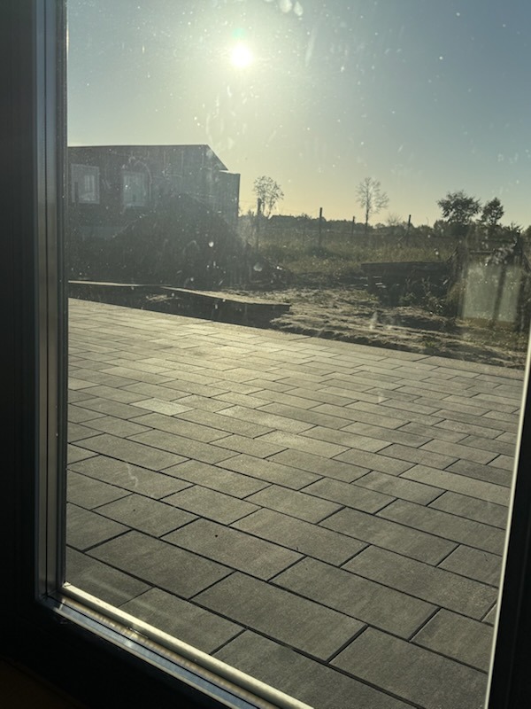 Ułożona, szara kostka brukowa widziana przez okno. W tle budynek w budowie i drzewa. Słoneczny dzień.