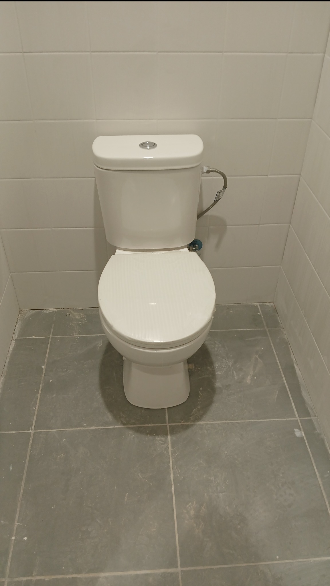 Biała toaleta kompaktowa po montażu w łazience wykończonej płytkami w Rumi. Widoczne przyłącze. Ujęcie z przodu, na podłodze szare płytki.