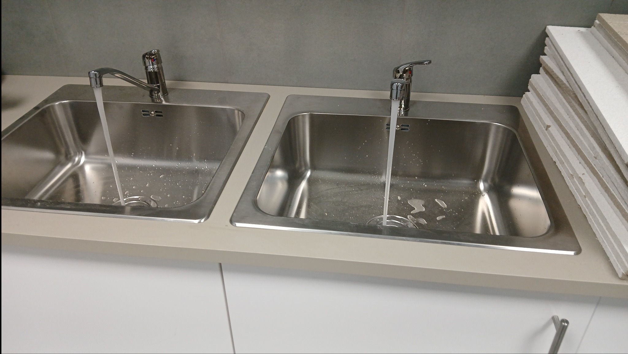 Dwa stalowe zlewozmywaki z kranami, z których leci woda. Obok ułożone płyty. Widok z góry, oświetlenie neutralne, wnętrze w trakcie wykańczania.