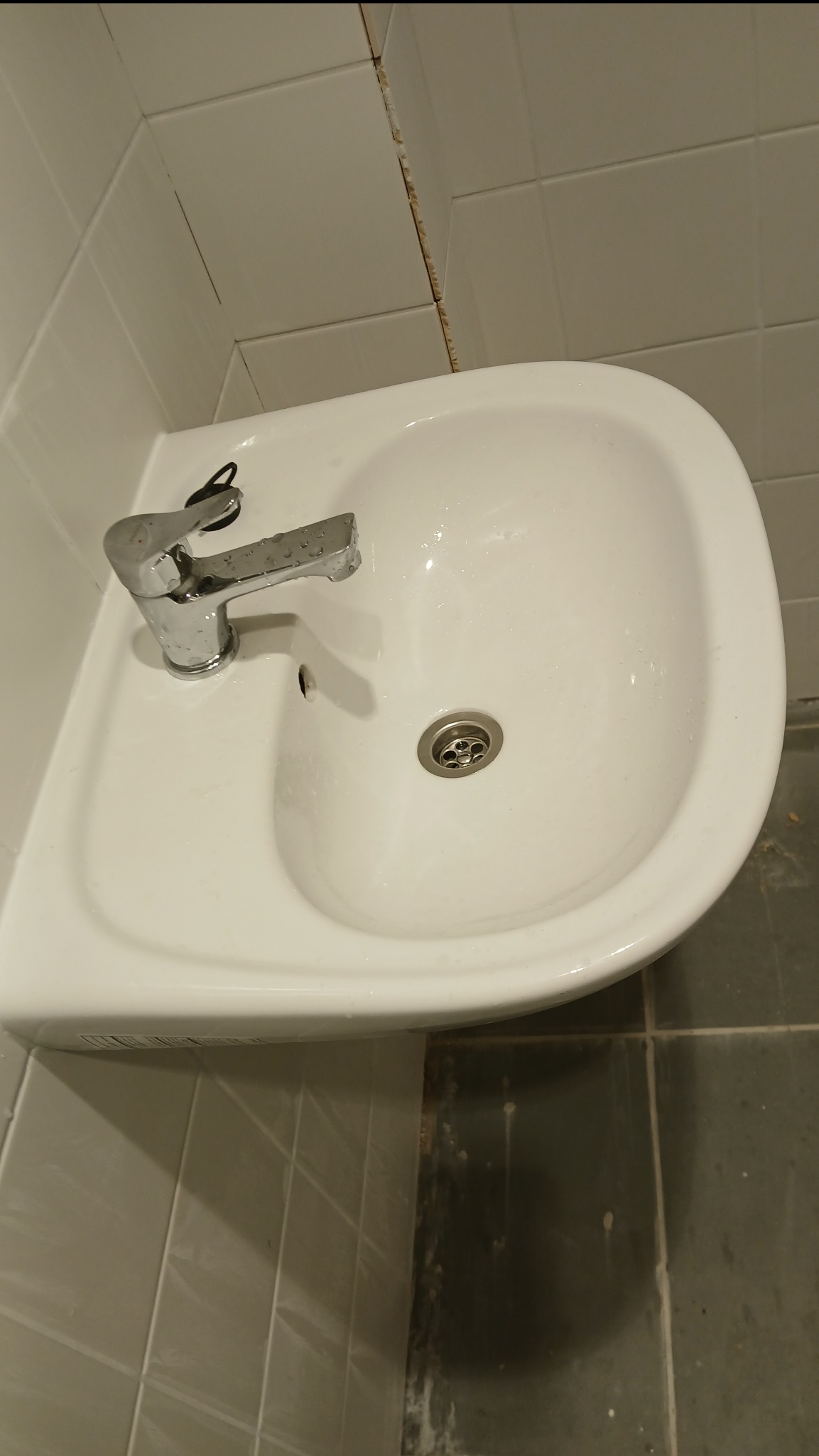 Biała umywalka z chromowaną baterią i odpływem, zamontowana w rogu łazienki z białymi płytkami. Widoczne ślady po remoncie na fugach i podłodze.