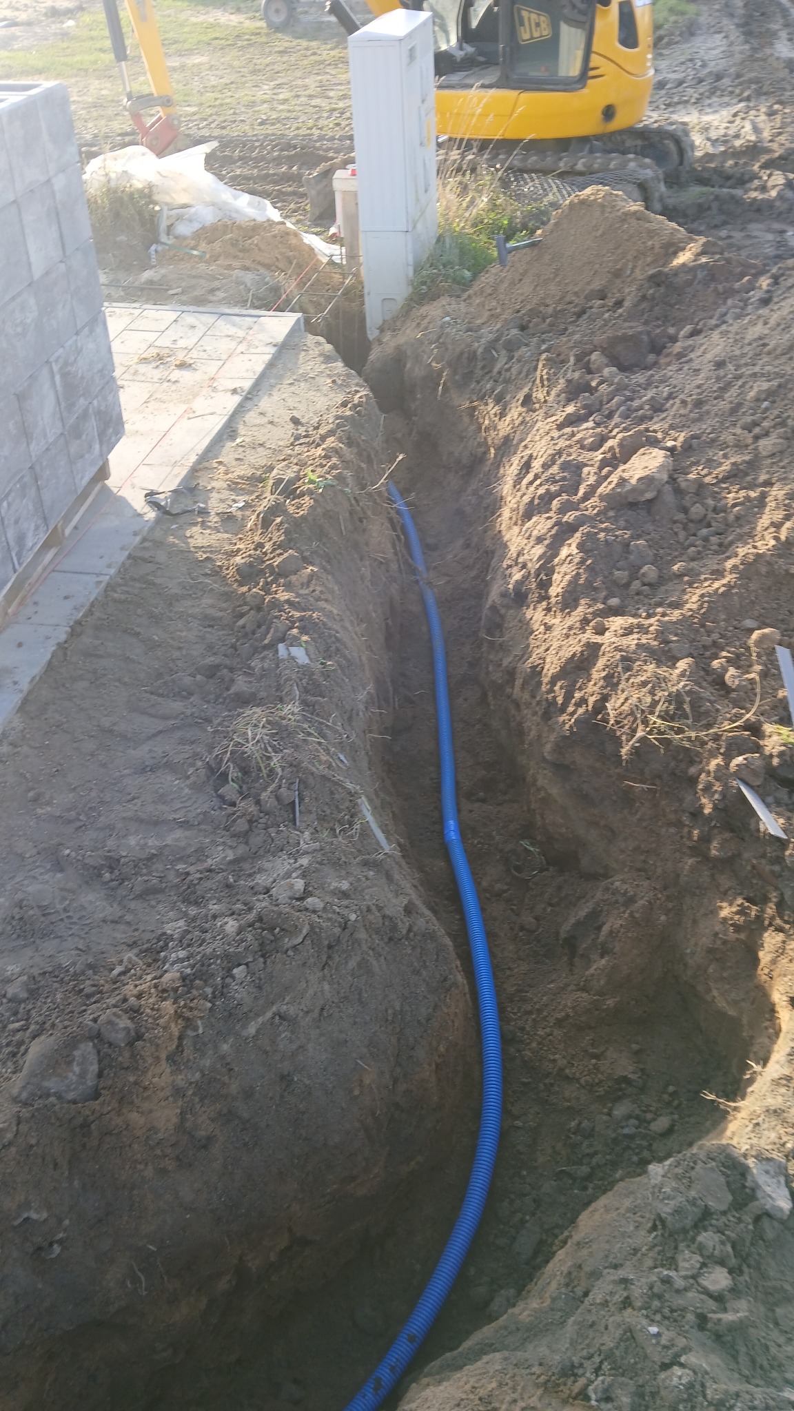 Niebieska rura w wykopie ziemnym, prawdopodobnie pod instalację. W tle widoczny sprzęt budowlany i elementy konstrukcyjne. Ujęcie z góry, w dziennym świetle.