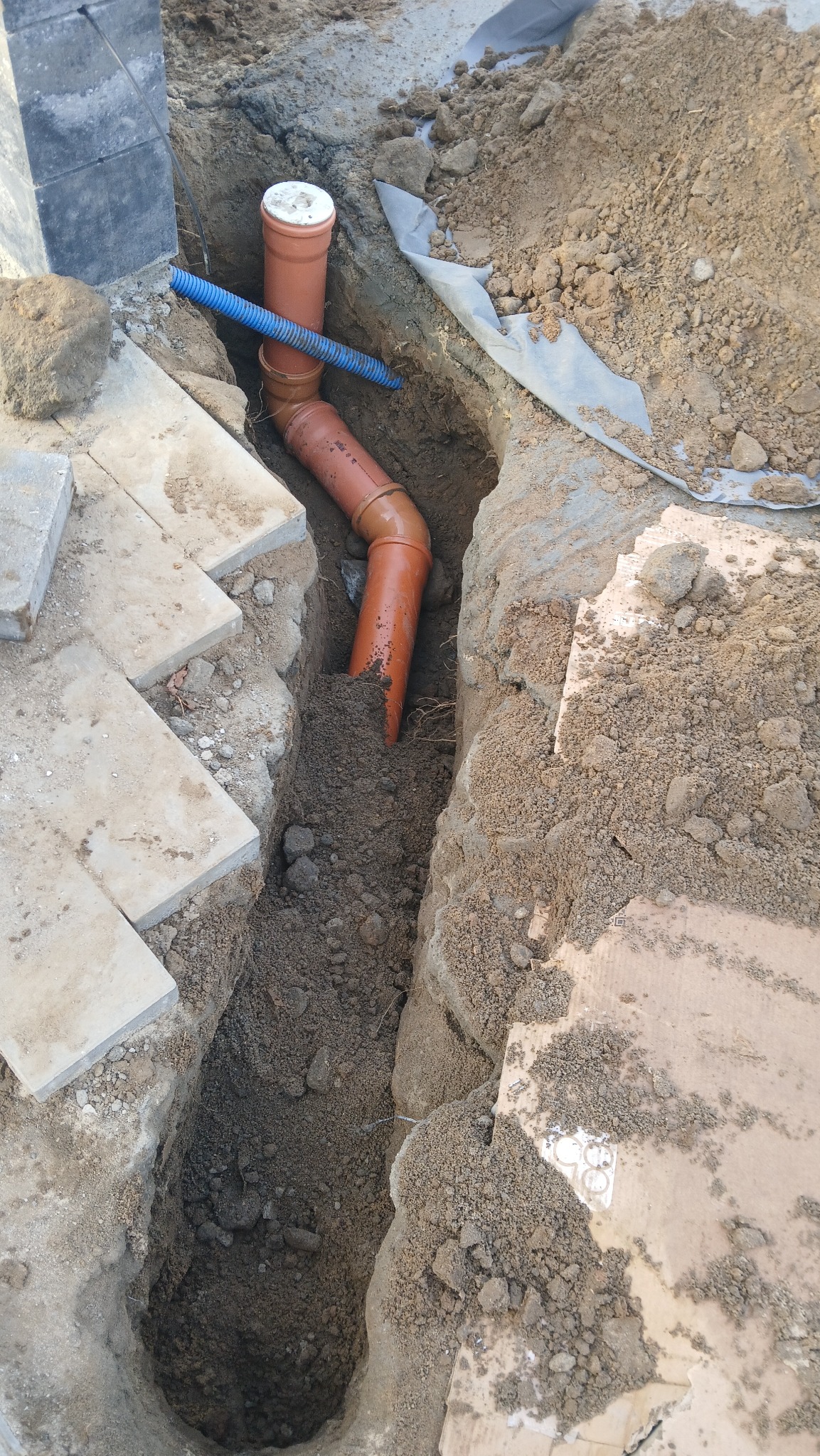 Wykop z ułożonymi rurami kanalizacyjnymi z tworzywa sztucznego, widoczne elementy kostki brukowej i ziemia. Rury w kolorze pomarańczowym i niebieskim.
