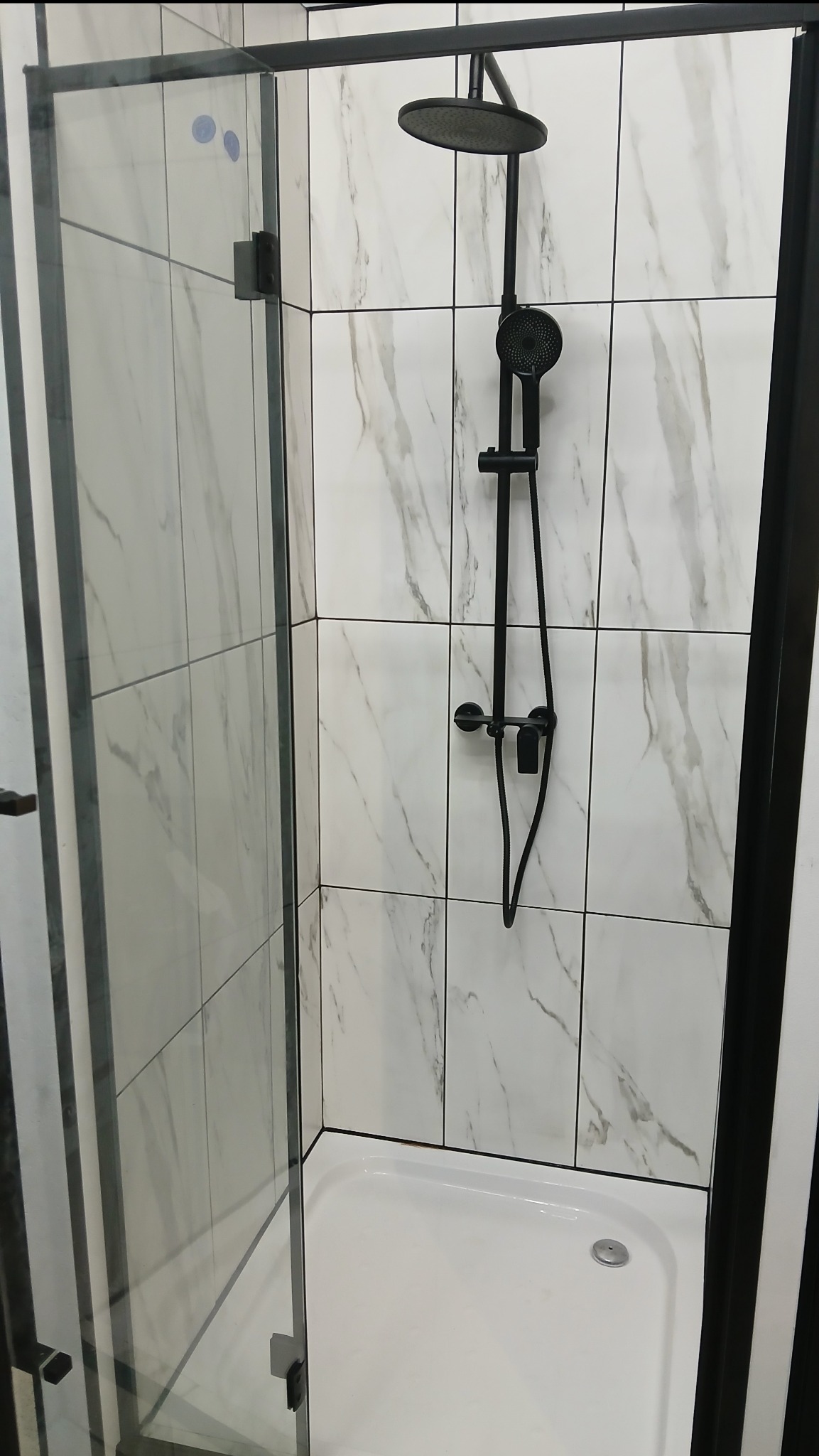 Nowoczesna kabina prysznicowa z czarną armaturą i białymi płytkami imitującymi marmur. Szklane drzwi z czarnymi zawiasami. Minimalistyczny design.