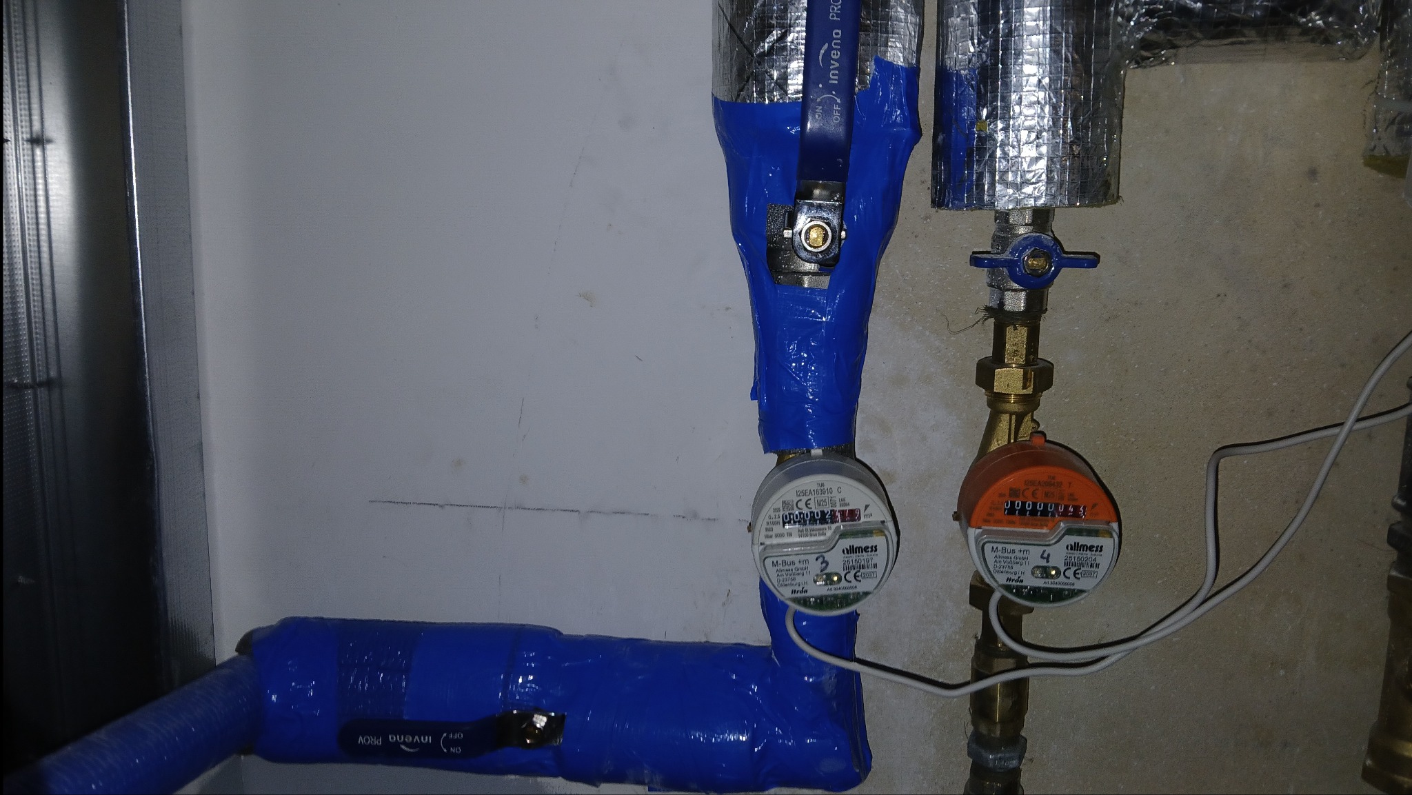 Dwa wodomierze, jeden owinięty niebieską taśmą, drugi z mosiężnymi rurami, zamontowane na ścianie. Widoczne zawory i izolacja termiczna.