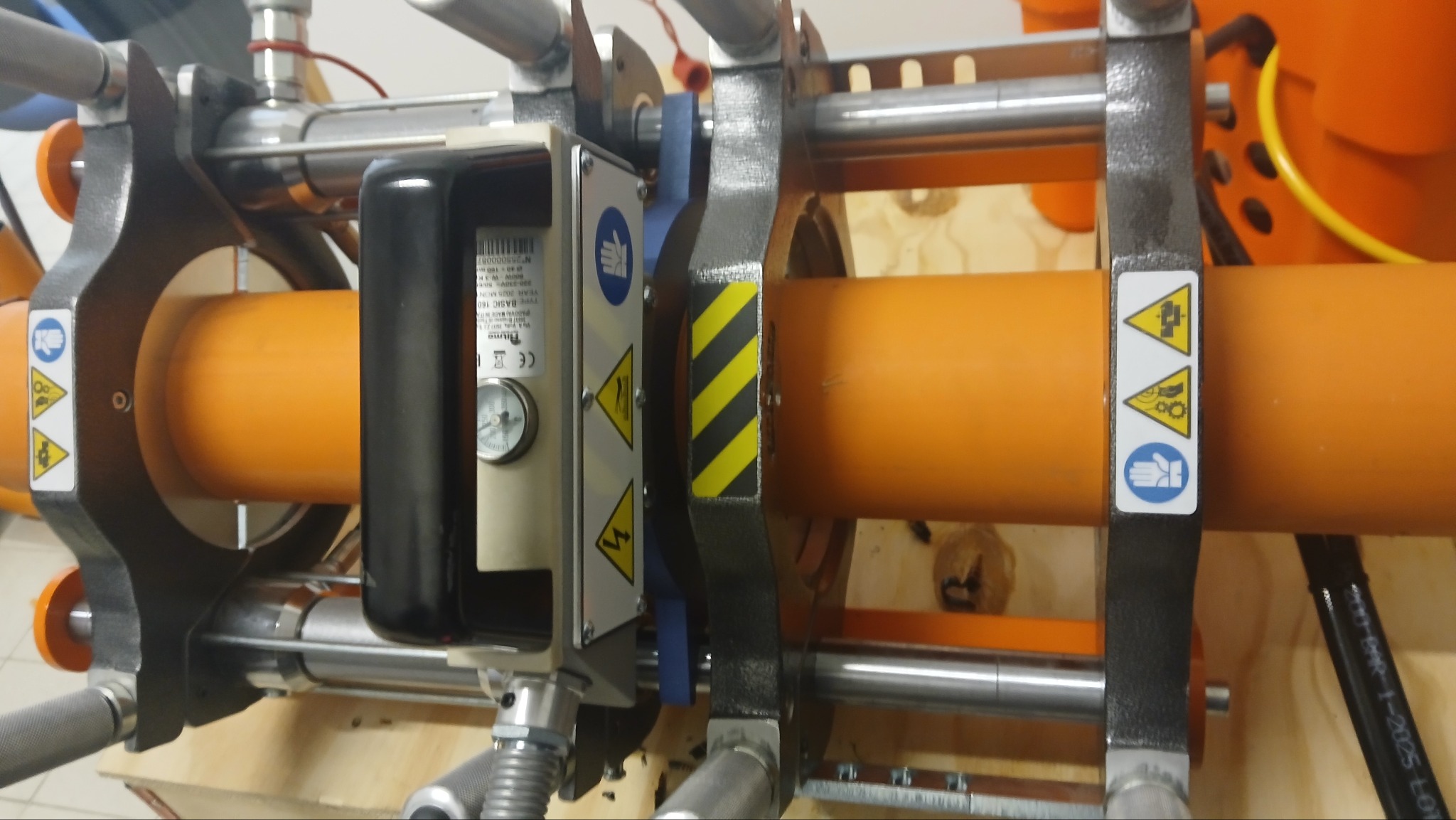 Zgrzewarka doczołowa do rur z tworzyw sztucznych, pomarańczowa rura w uchwycie, widoczne oznaczenia bezpieczeństwa i manometr. Sprzęt hydrauliczny w trakcie pracy.