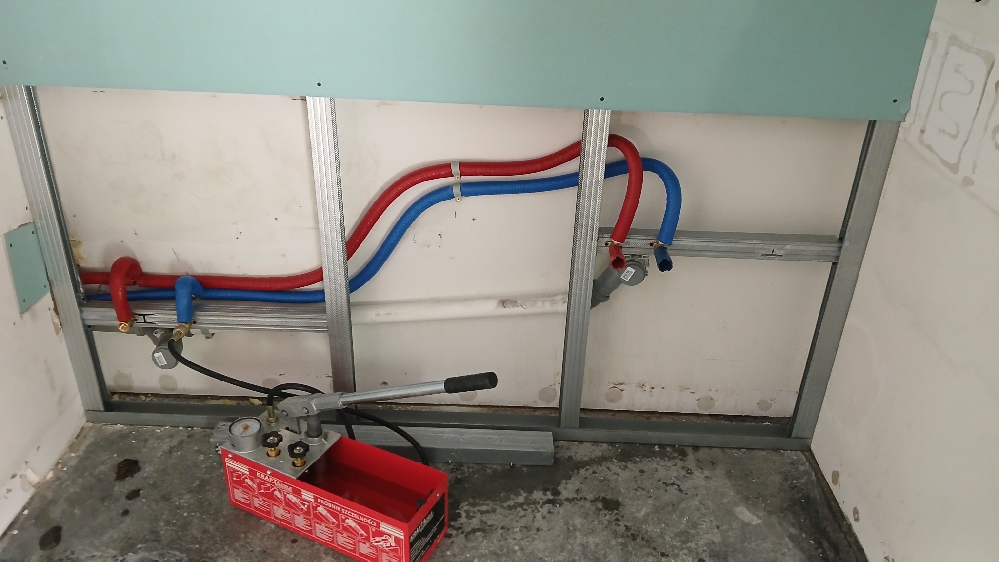 Instalacja wodna z rurami PEX (czerwona i niebieska) zamontowanymi na metalowym stelażu, widoczny manometr i pompa ciśnieniowa do testu szczelności.