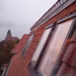 Czerwony dach z oknami dachowymi i widokiem na miasto w tle. Widoczna tekstura dachówek i ramy okien. Pochmurne niebo, perspektywa z dołu.