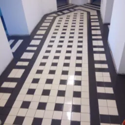 Wzorzysta, czarno-biała podłoga z płytek w korytarzu, układana w pasy i kwadraty. Widoczny fragment mopa w prawym dolnym rogu kadru. Białe ściany.