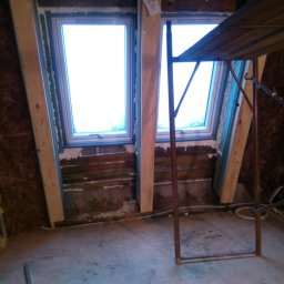 MAR-DOM Usługi remontowo-budowlane - Montaż okien dachowych w skośnym dachu z widocznymi drewnianymi belkami. Etap prac z użyciem pianki montażowej i rusztowania. Widok na niebo przez okna.