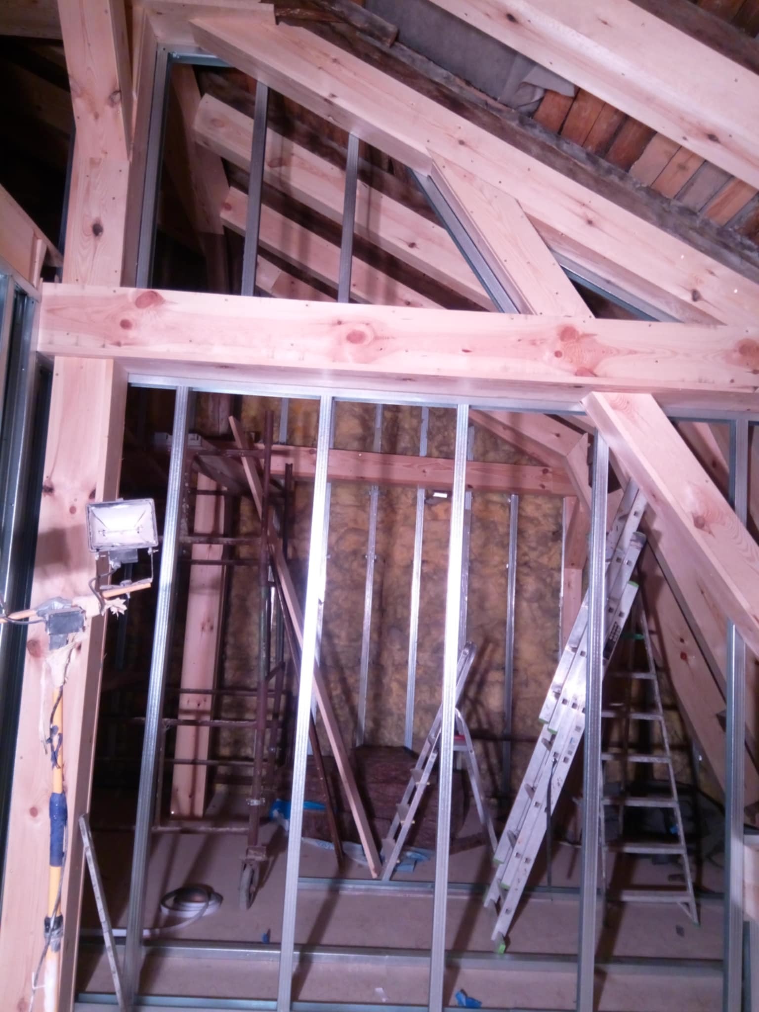 Konstrukcja ścianki działowej z profili stalowych w surowym wnętrzu poddasza z drewnianą więźbą dachową. Widoczne drabiny i oświetlenie robocze.