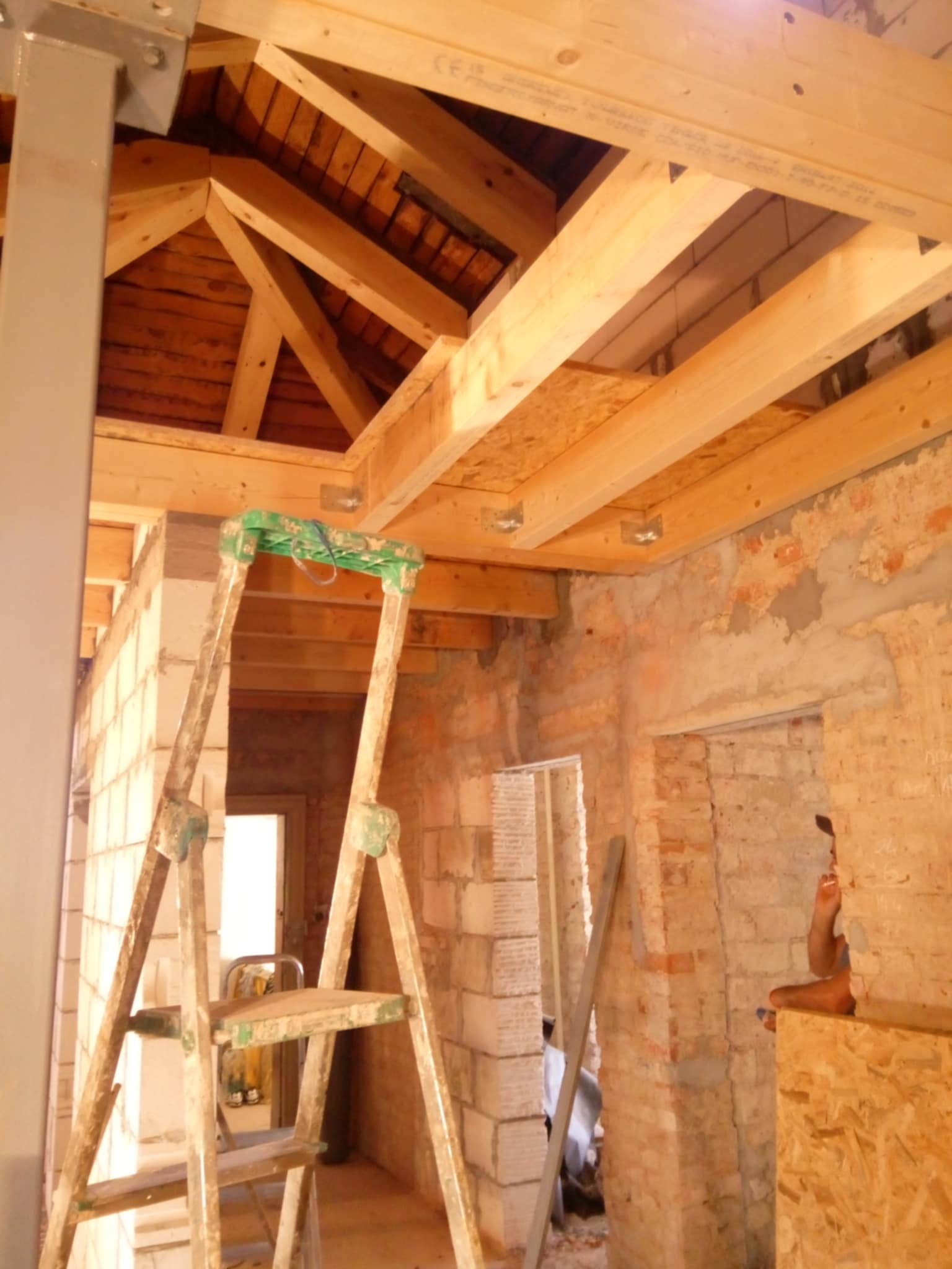 Wnętrze w trakcie remontu: drewniana konstrukcja stropu, surowe ściany z cegły i bloczków, drabina malarska. Widoczne ślady prac budowlanych.