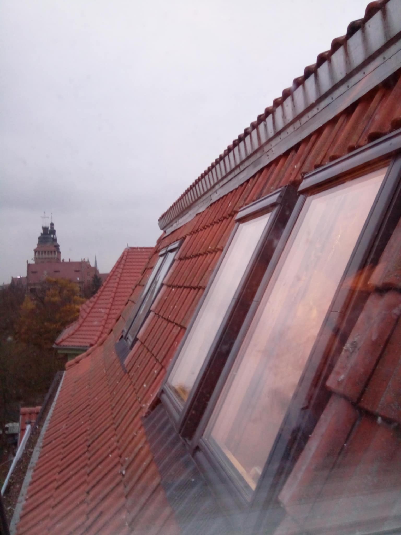 Czerwony dach z oknami dachowymi i widokiem na miasto w tle. Widoczna tekstura dachówek i ramy okien. Pochmurne niebo, perspektywa z dołu.