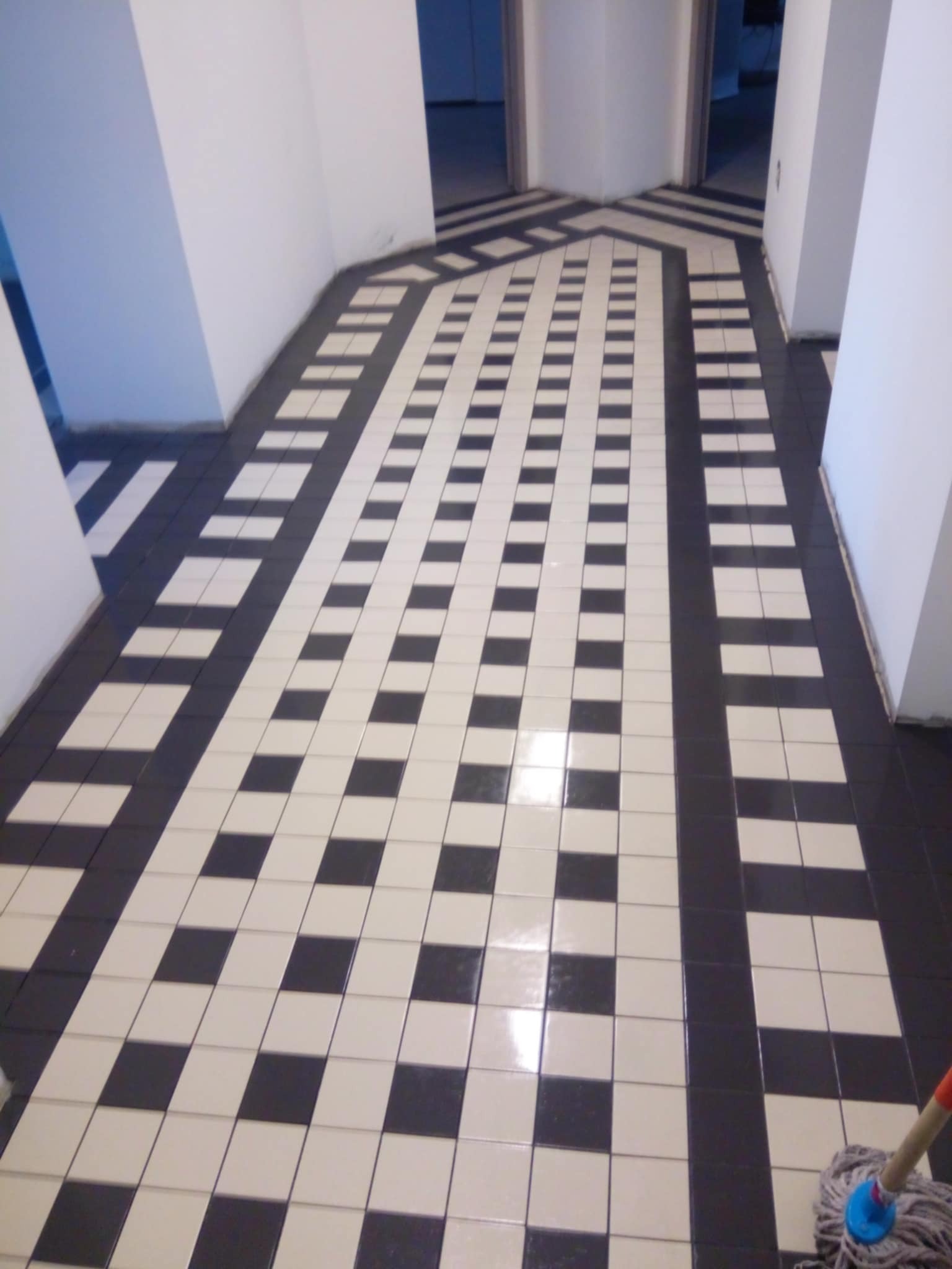 Wzorzysta, czarno-biała podłoga z płytek w korytarzu, układana w pasy i kwadraty. Widoczny fragment mopa w prawym dolnym rogu kadru. Białe ściany.