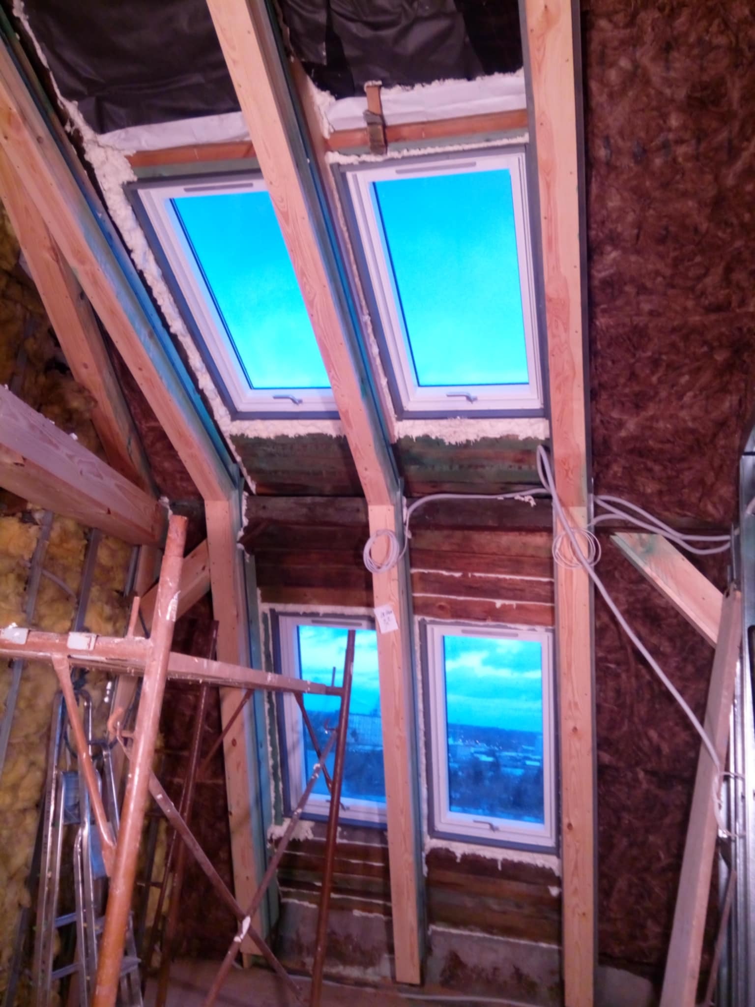 Adaptacja poddasza: montaż okien dachowych z drewnianą konstrukcją, ocieplenie wełną mineralną, widoczne rusztowania. Prace wykończeniowe w toku.