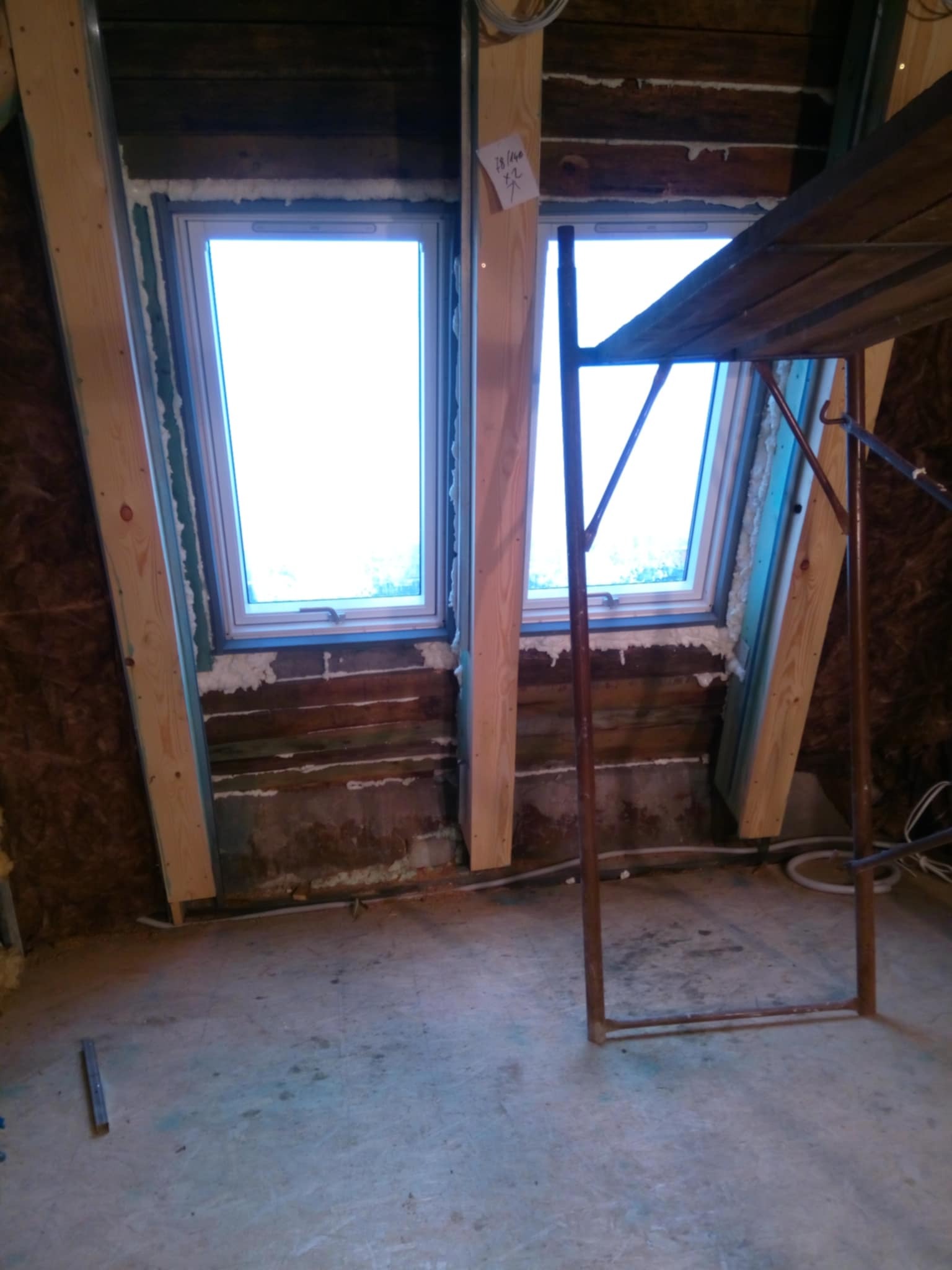 Montaż okien dachowych w skośnym dachu z widocznymi drewnianymi belkami. Etap prac z użyciem pianki montażowej i rusztowania. Widok na niebo przez okna.