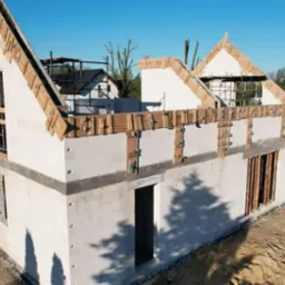 Budowa domu z białych bloczków, widoczne drewniane szalunki przy dachu i oknach. W tle las i częściowo widoczny inny budynek.