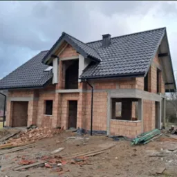 Surowy dom z czerwonej cegły z czarnym dachem w trakcie budowy, widoczne niezabudowane otwory okienne i drzwiowe, teren budowy z gruzem i drewnem.