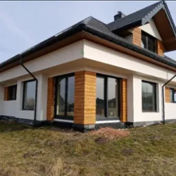 Nowoczesny dom z jasną elewacją, czarnymi oknami i drewnianymi akcentami. Widoczne orynnowanie i fragment dachu. Otoczenie: trawnik i elementy krajobrazu.