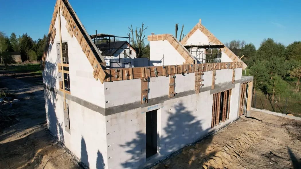 Budowa domu z białych bloczków, widoczne drewniane szalunki przy dachu i oknach. W tle las i częściowo widoczny inny budynek.