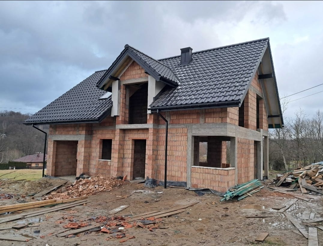 Surowy dom z czerwonej cegły z czarnym dachem w trakcie budowy, widoczne niezabudowane otwory okienne i drzwiowe, teren budowy z gruzem i drewnem.