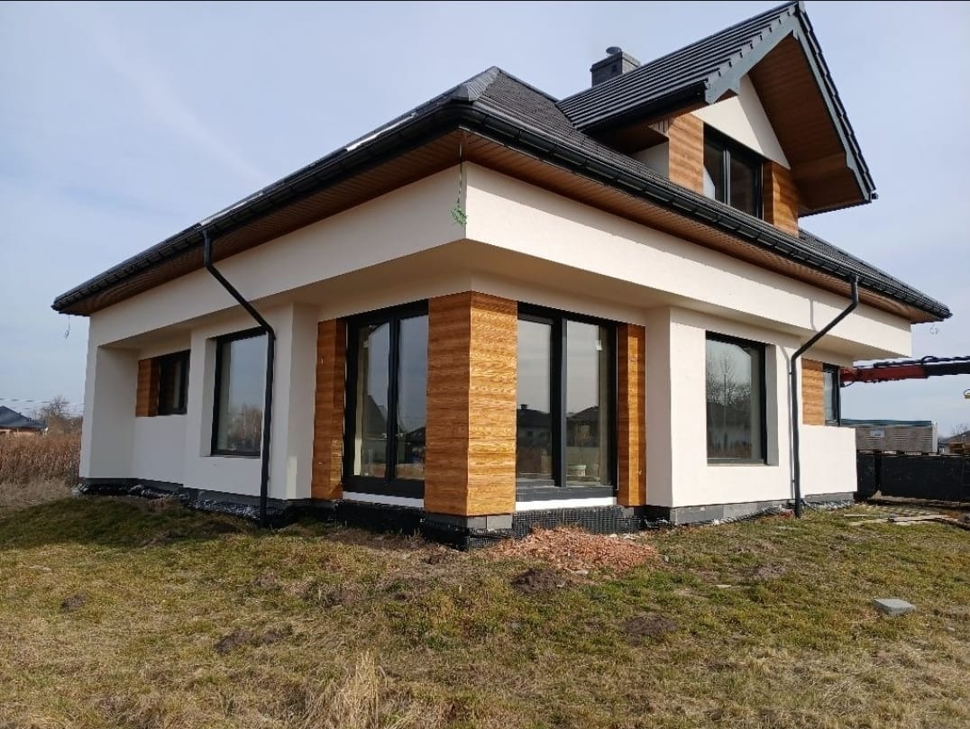 Nowoczesny dom z jasną elewacją, czarnymi oknami i drewnianymi akcentami. Widoczne orynnowanie i fragment dachu. Otoczenie: trawnik i elementy krajobrazu.