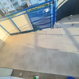 Wykończony balkon z jasnymi płytkami i niebieską barierką, widoczne elementy montażowe płytek, światło słoneczne. Widok z góry.