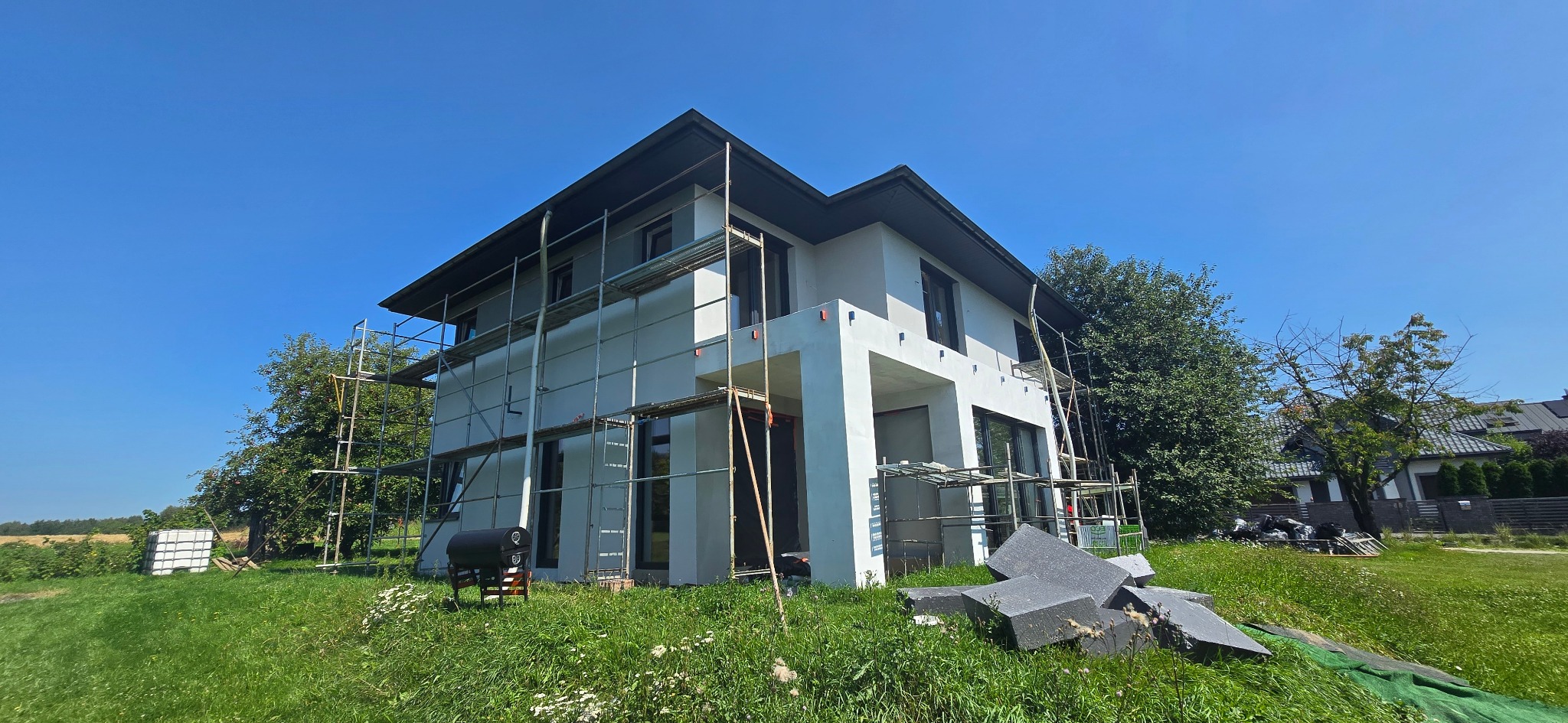 Dwukondygnacyjny dom w trakcie ocieplania, z widocznymi rusztowaniami i stertą grafitowego styropianu na trawniku, błękitne niebo w tle.