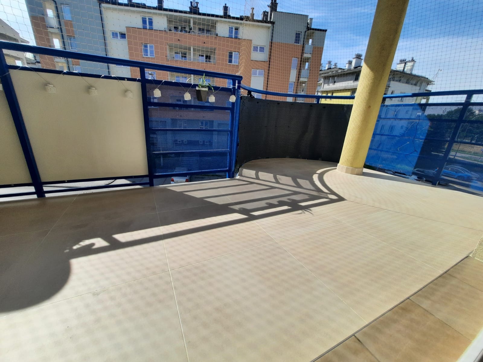 Wykończony balkon z beżowymi płytkami, niebieską balustradą i osłoną. Na balustradzie wiszą doniczki z roślinami. Widok na budynki w tle.