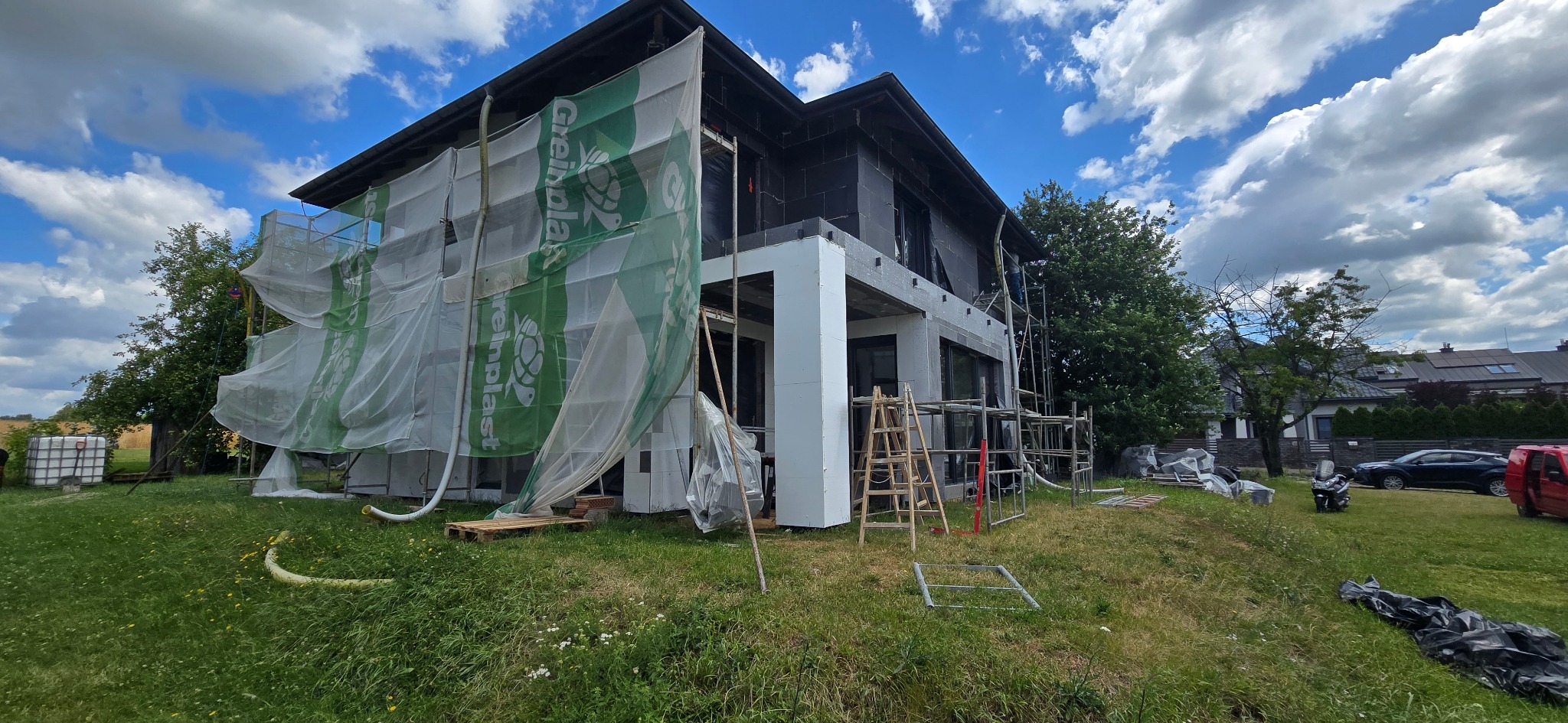 Dom w trakcie ocieplania styropianem, z rusztowaniami i siatką ochronną, widok z zewnątrz w słoneczny dzień. Etap prac przed wykończeniem elewacji.
